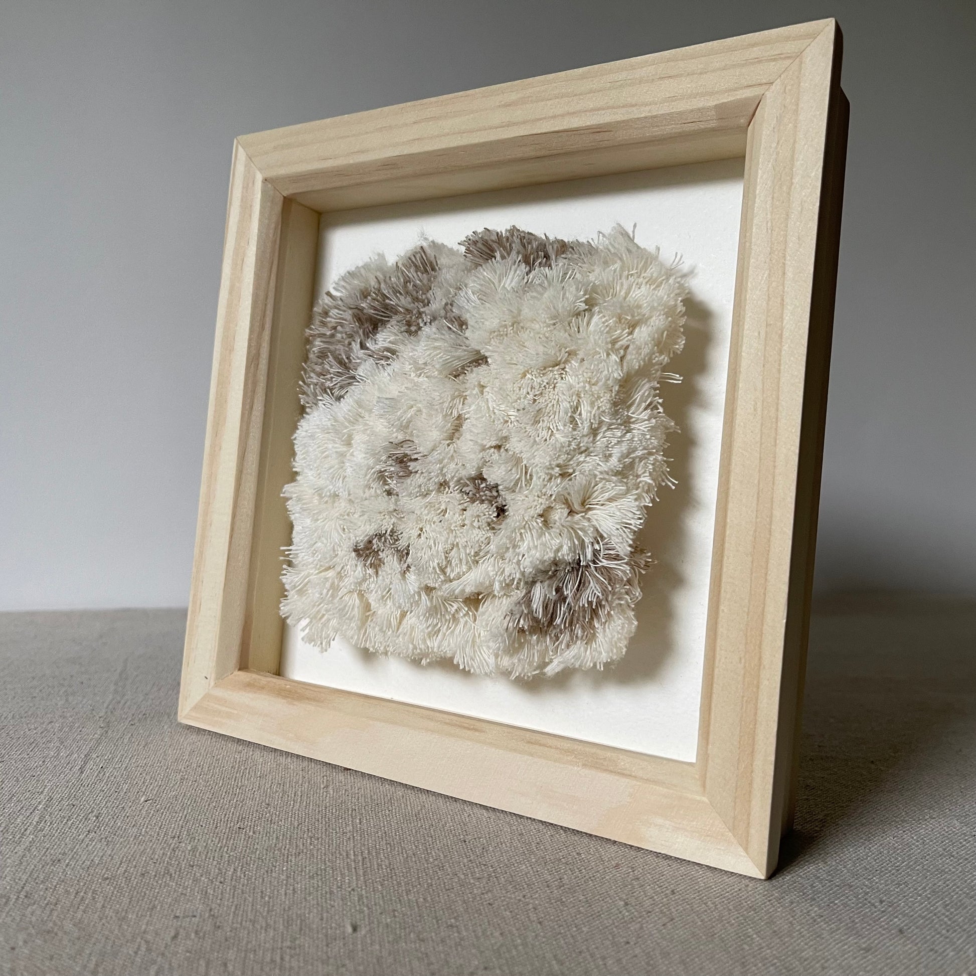Angled view of cream and beige fluffy woven textile art in square natural wood deep box frame