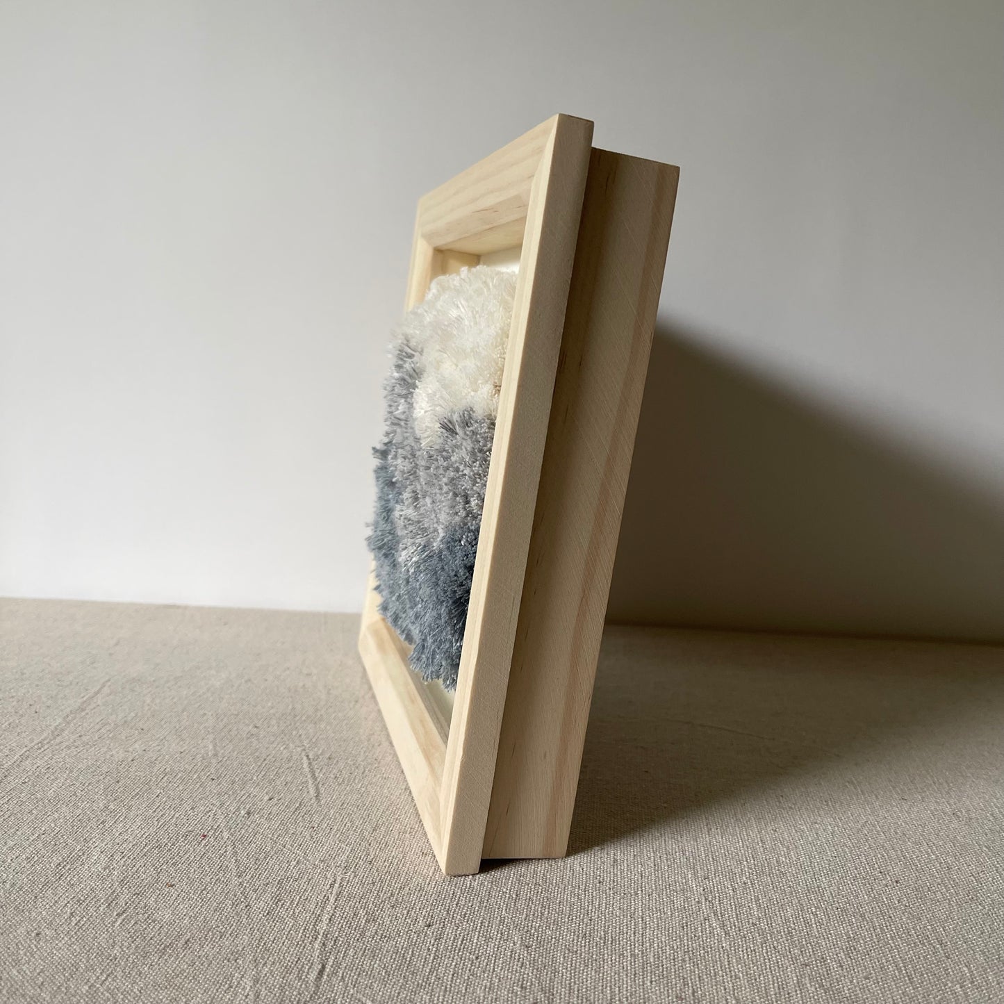 Side view of cream and blue fluffy textured woven textile art in square natural wood deep box frame