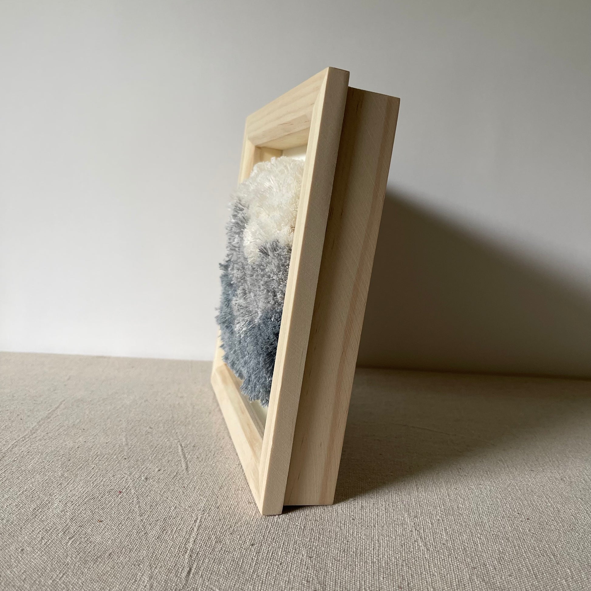 Side view of cream and blue fluffy textured woven textile art in square natural wood deep box frame