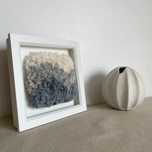 Cream and blue fluffy textured woven textile art in square white painted wood deep box frame on linen covered table next to scandi style vase