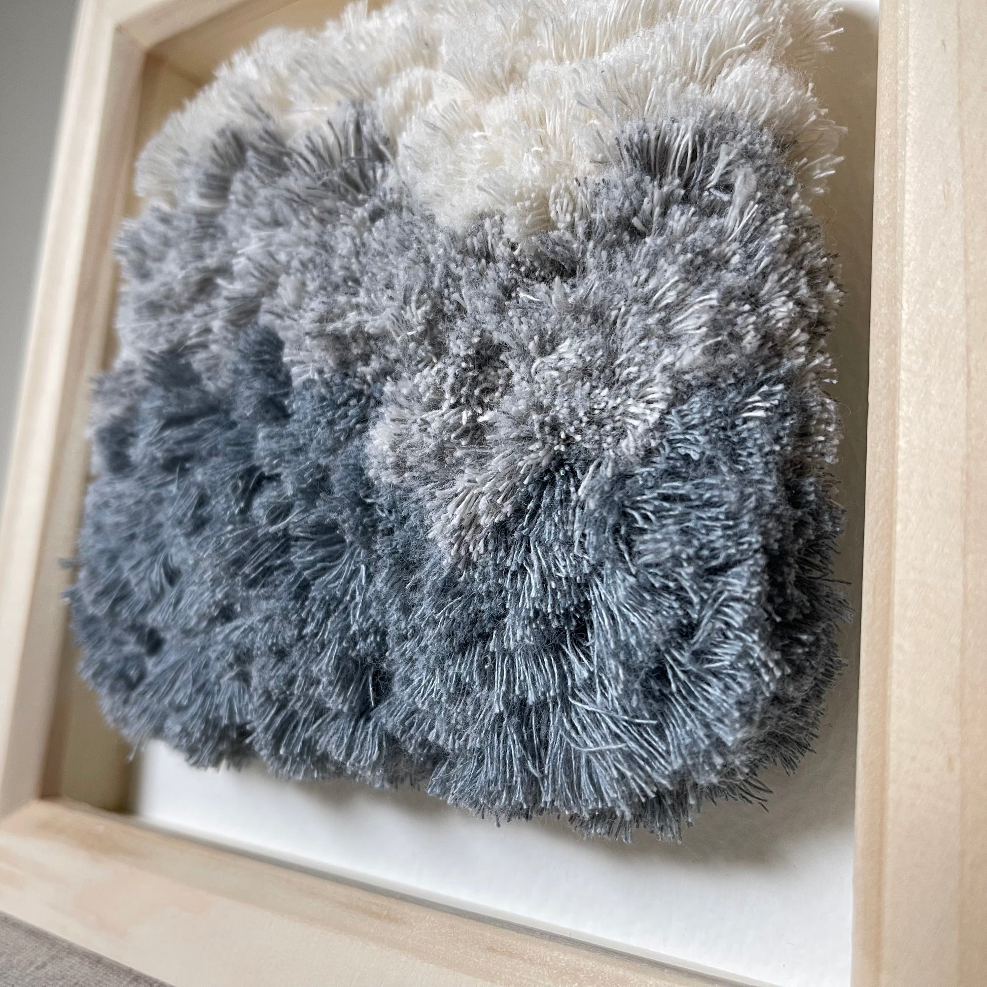 Close up view of cream and blue fluffy textured woven textile art in square natural wood frame