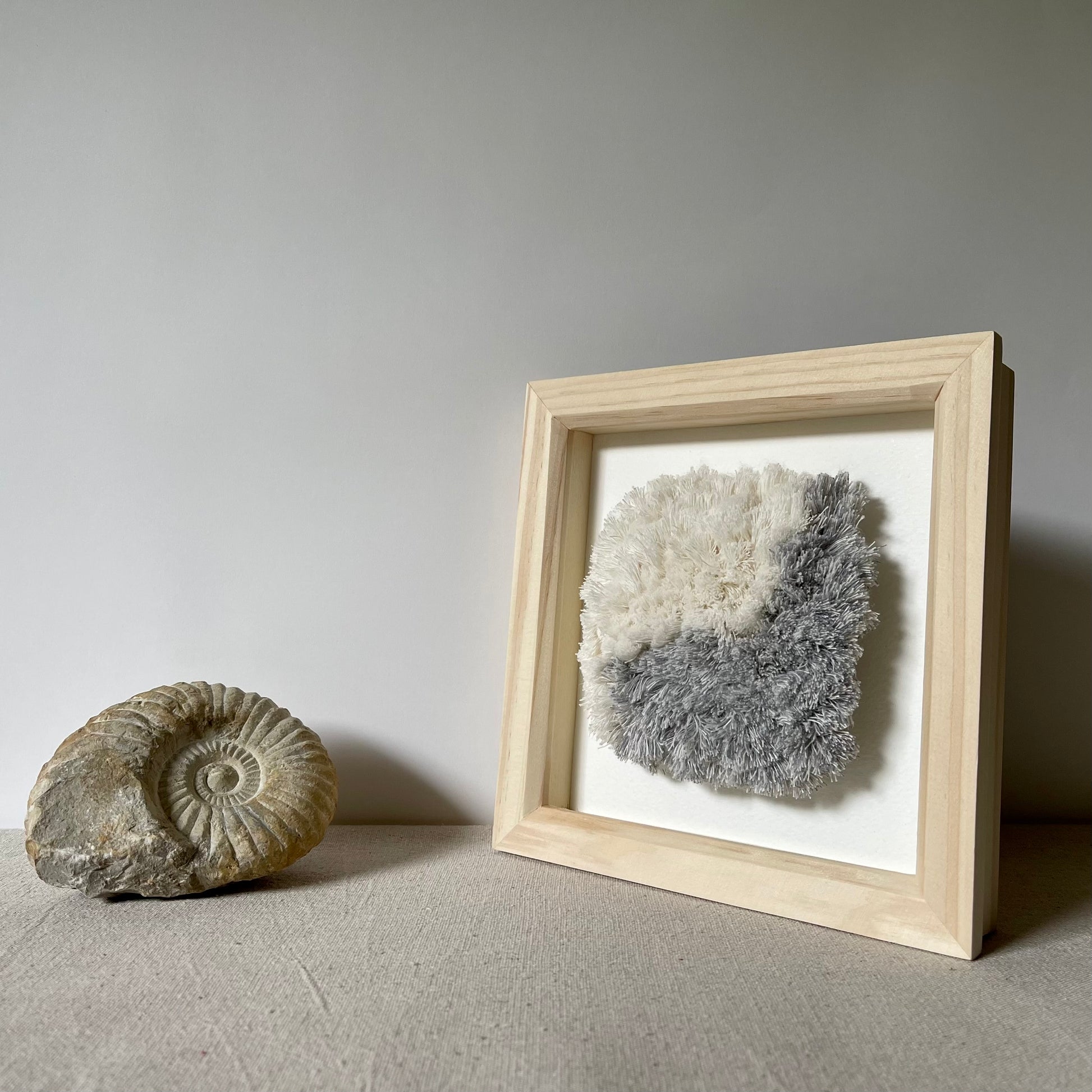 Blue and cream fluffy woven textile art in square natural wood box frame next to ammonite