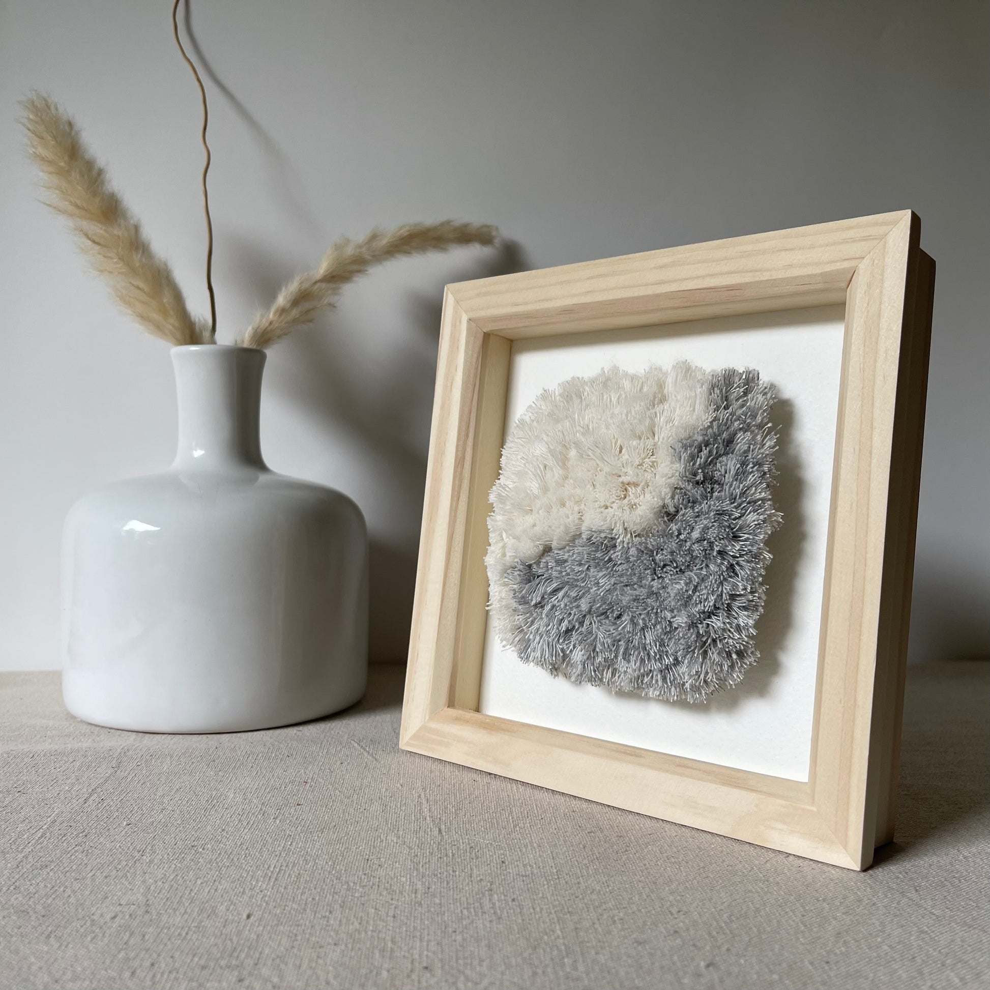Blue and cream fluffy textured woven textile art in square natural wood deep box frame next to white scandi vase