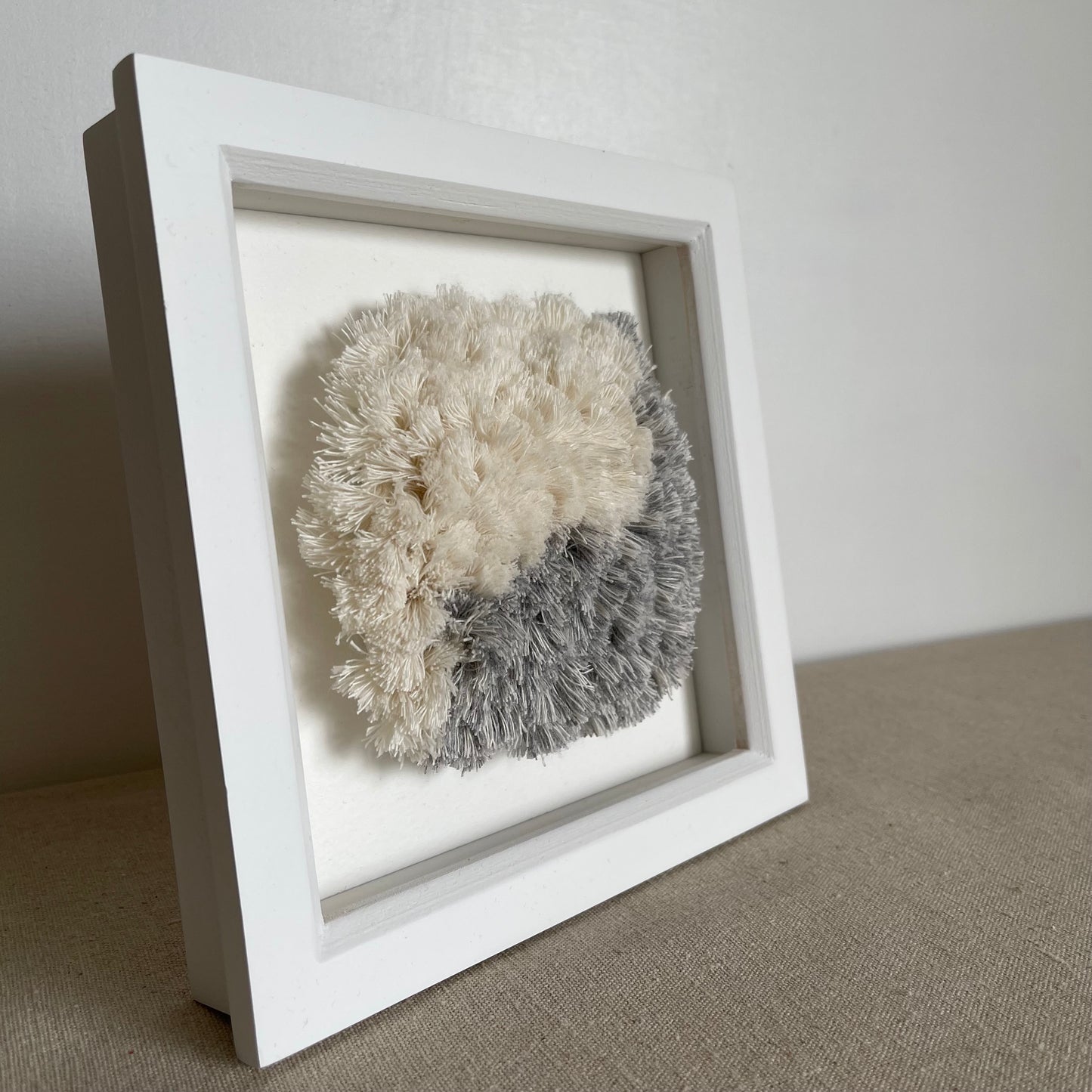 Square white painted wood deep box frame housing a blue and cream fluffy textured woven textile art