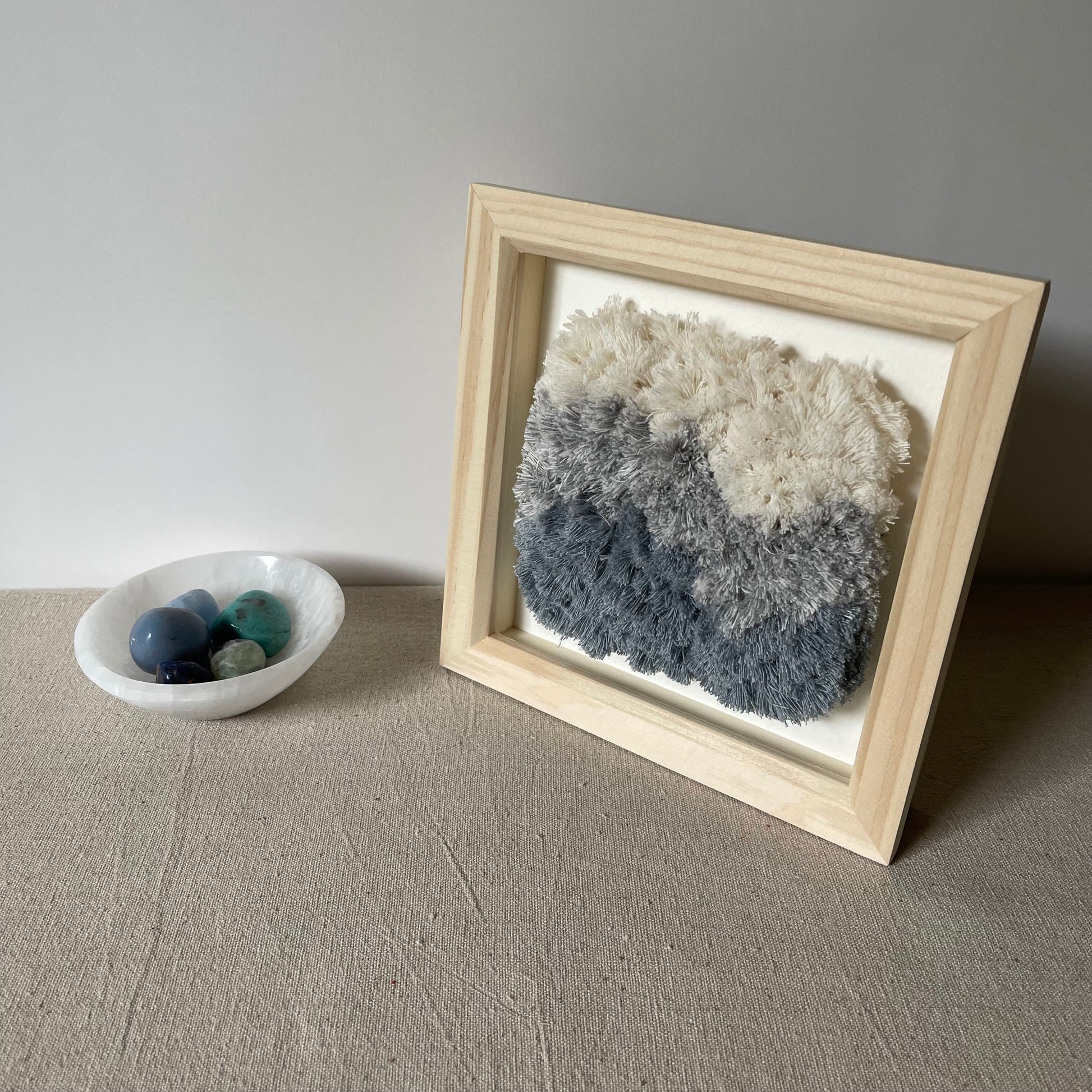 Cream and blue fluffy textured woven textile art in square natural wood deep box frame next to selenite bowl containing several blue crystals