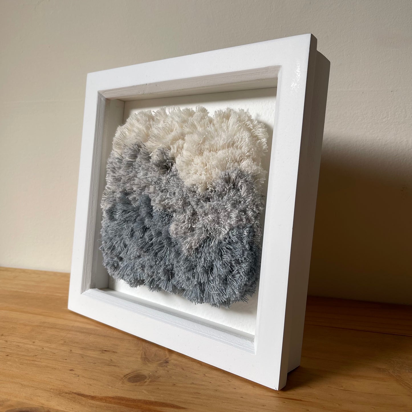 Angled view of cream and blue fluffy textured woven textile art in square white painted wood deep box frame