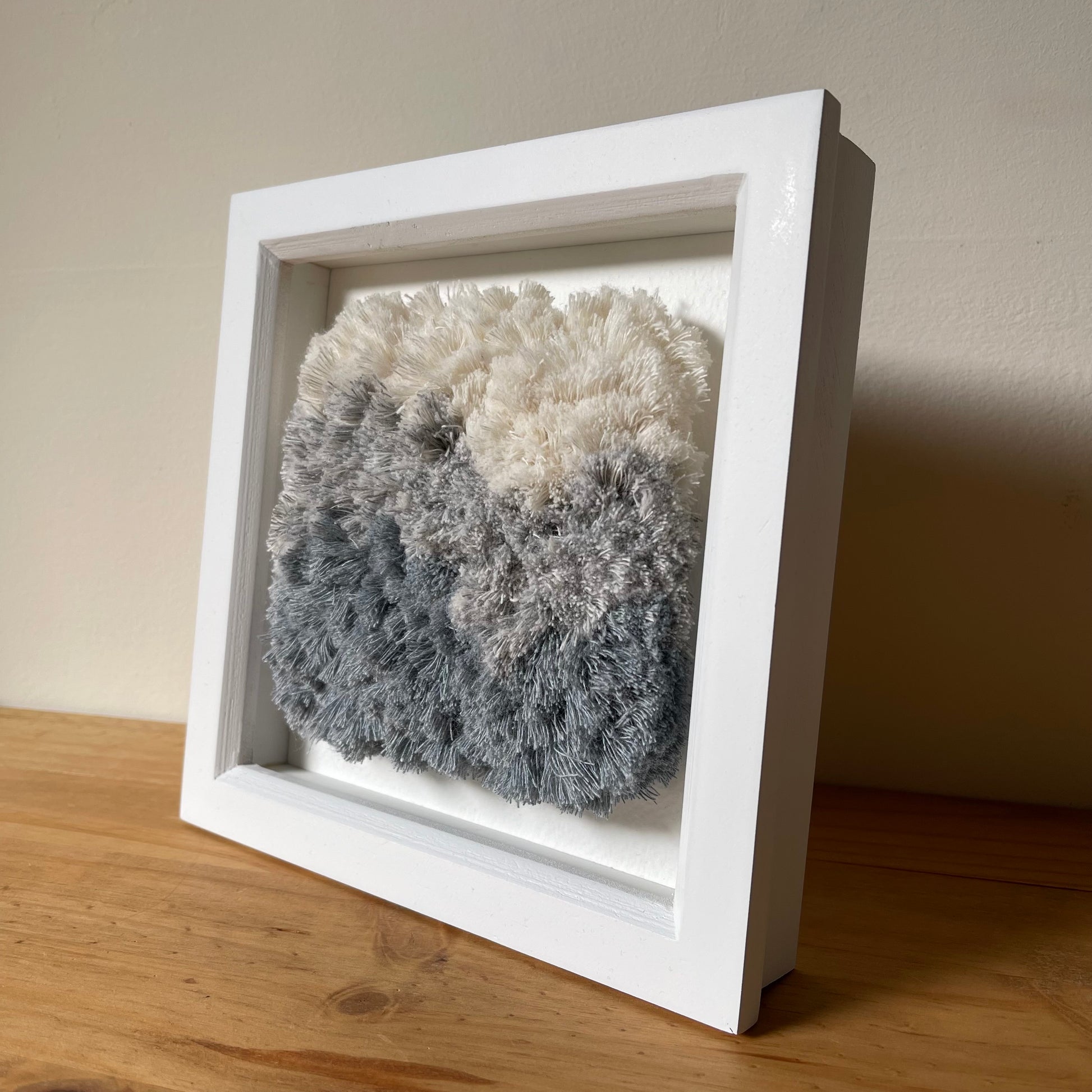 Angled view of cream and blue fluffy textured woven textile art in square white painted wood deep box frame