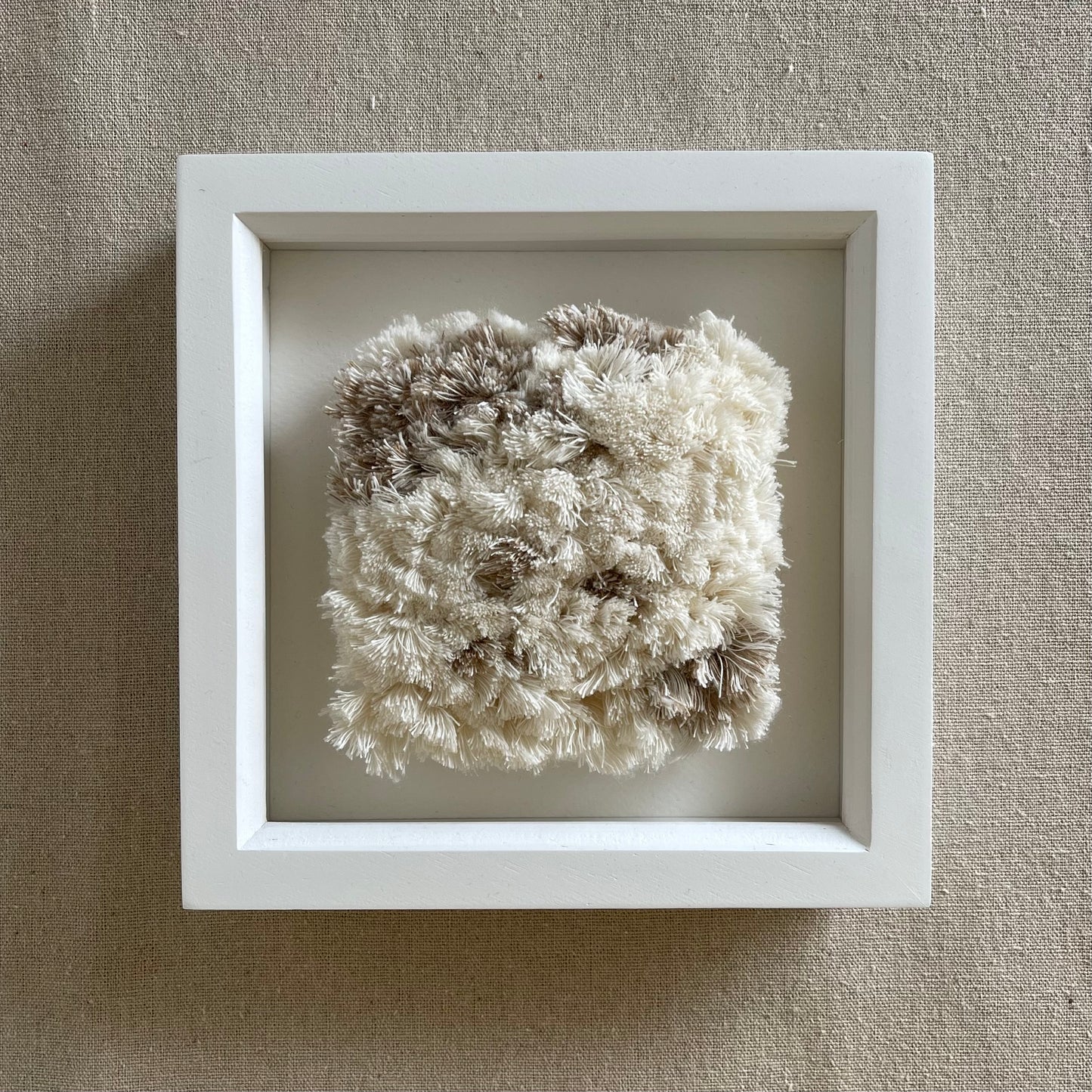 Front view of cream and beige fluffy textured woven textile art in square white painted wood deep box frame