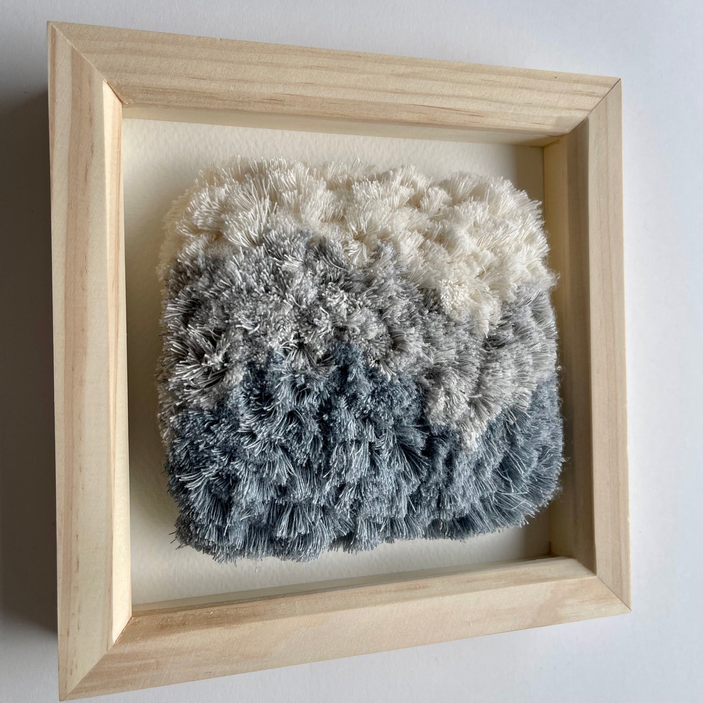 Angled view of cream and blue fluffy textured woven textile art in square natural wood deep box frame on wall
