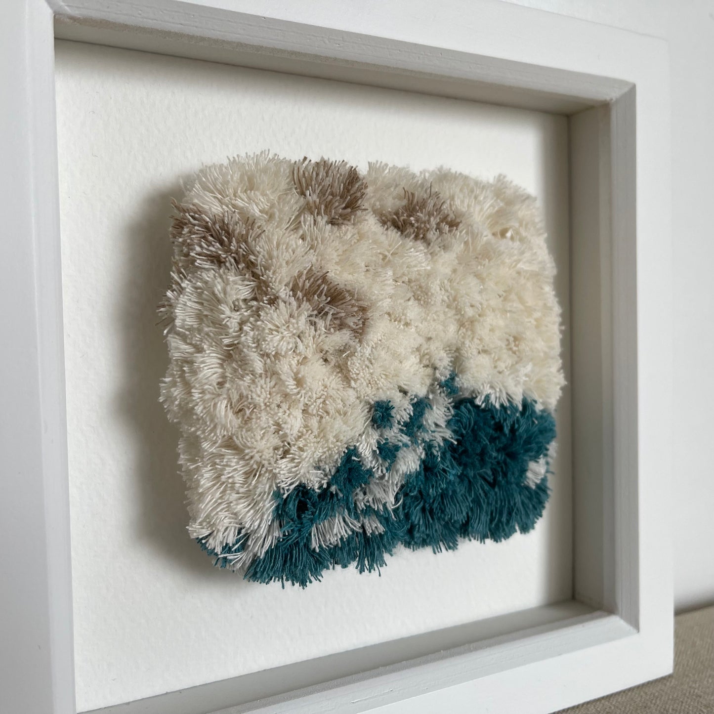 Side view of ream and ocean teal woven fluffy textured art in a square white painted wood deep box frame