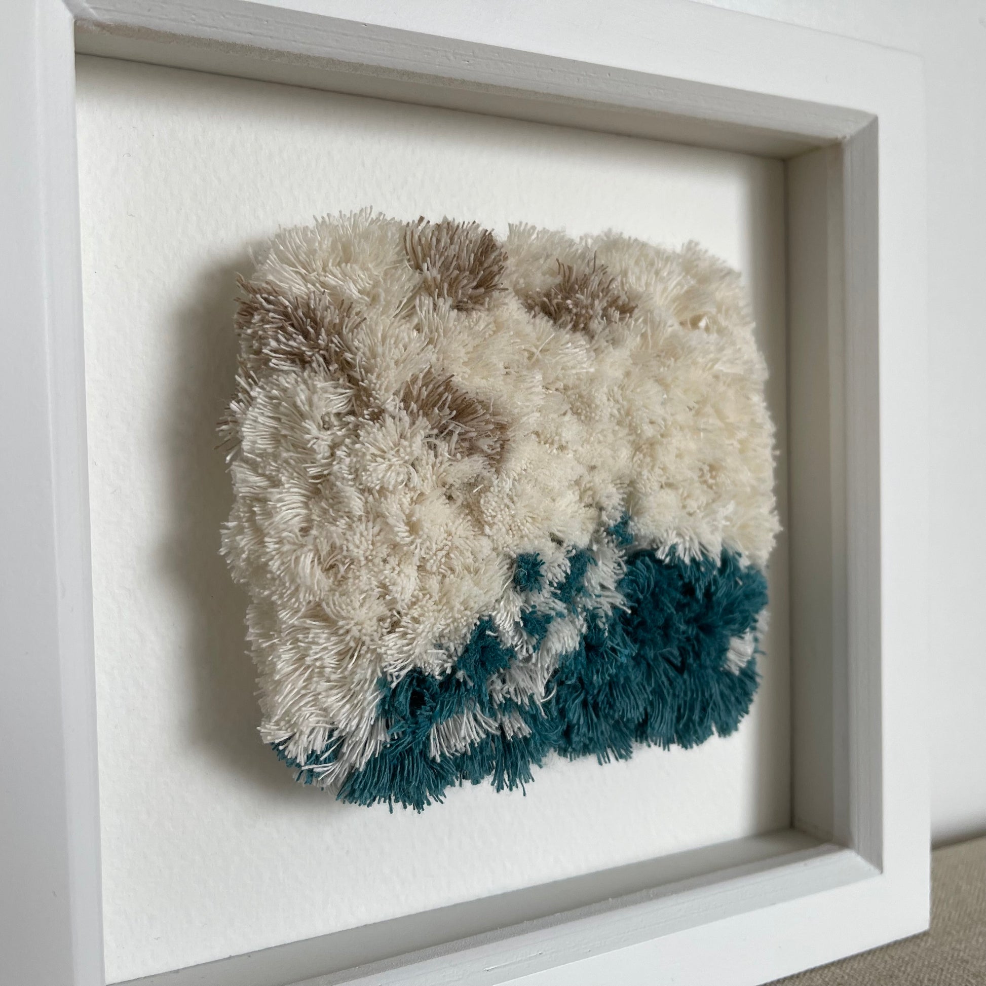 Side view of ream and ocean teal woven fluffy textured art in a square white painted wood deep box frame