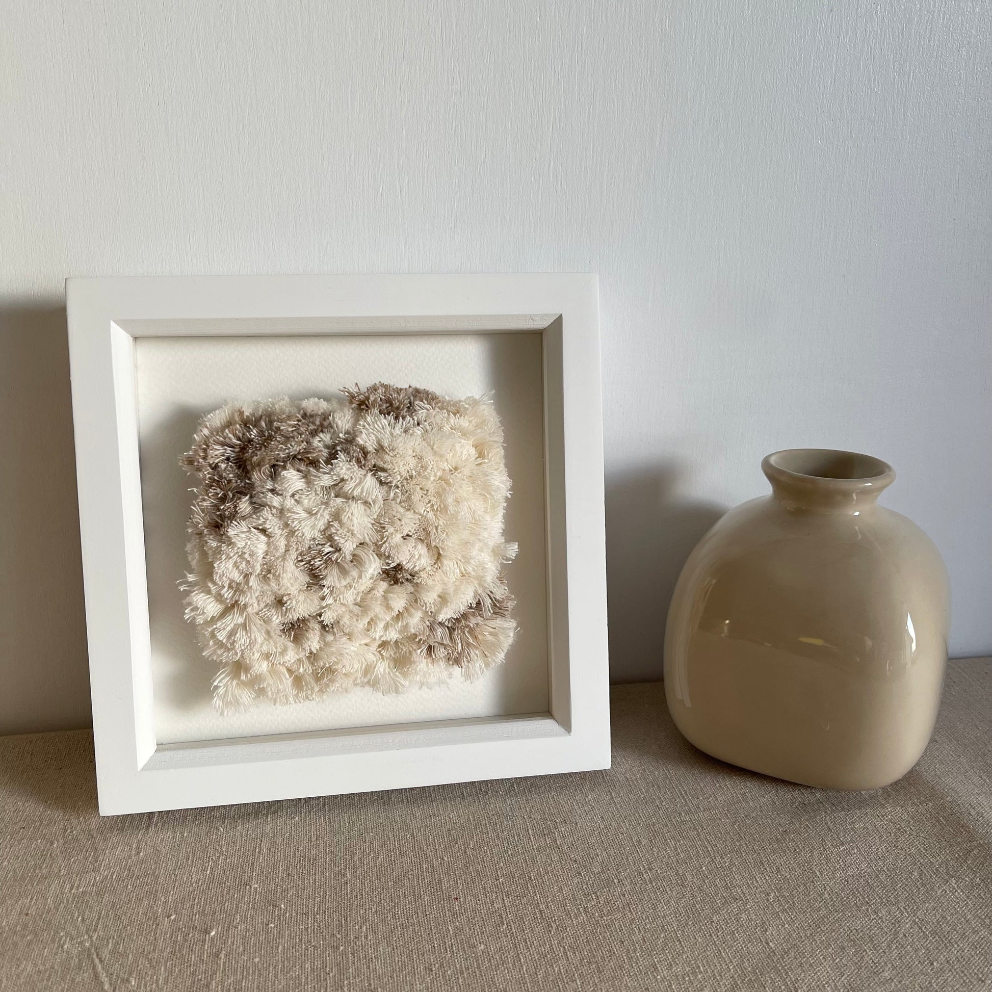 Cream and beige fluffy textured woven textile art in square white painted wood deep box frame next to scandi style vase