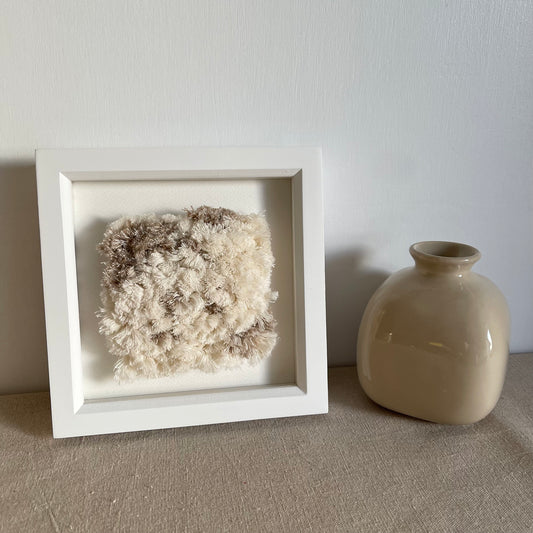 Cream and beige fluffy textured woven textile art in square white painted wood deep box frame next to scandi style vase