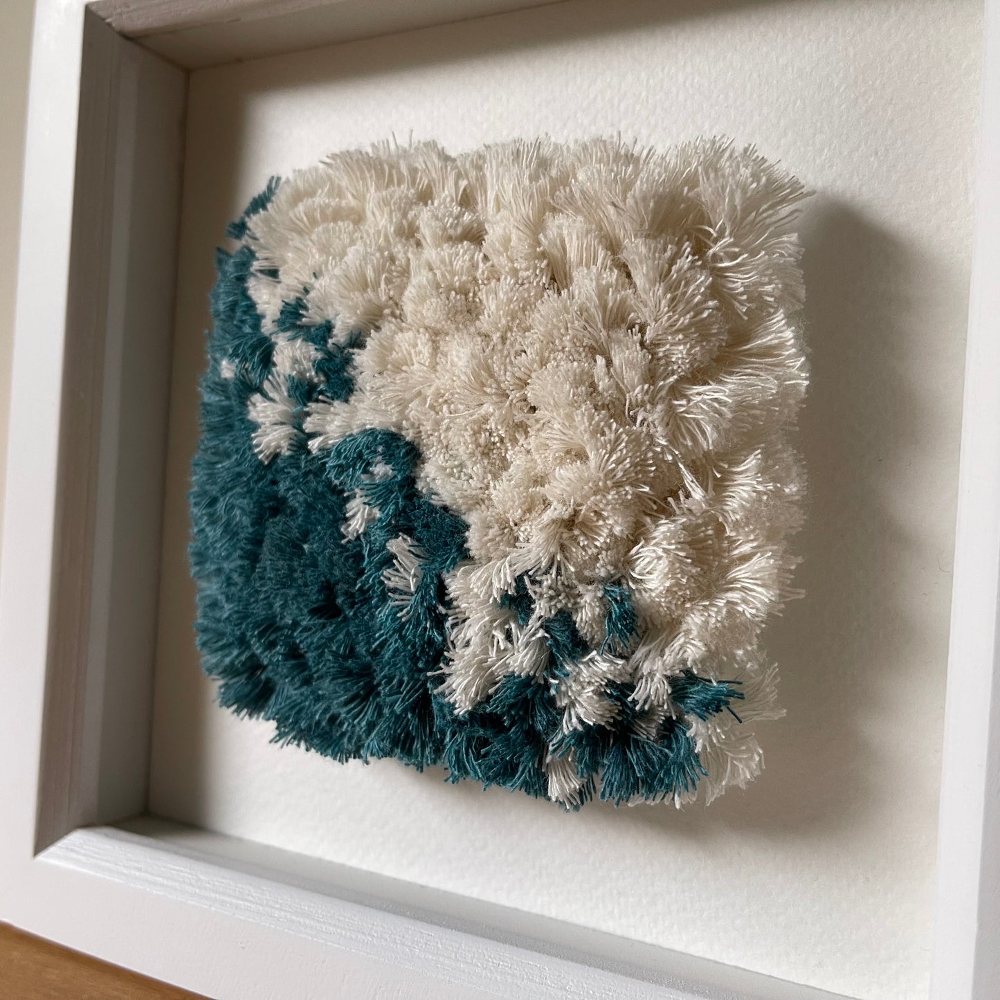 Angled close up view of teal and cream fluffy textured woven textile art in a square white painted wood deep box frame