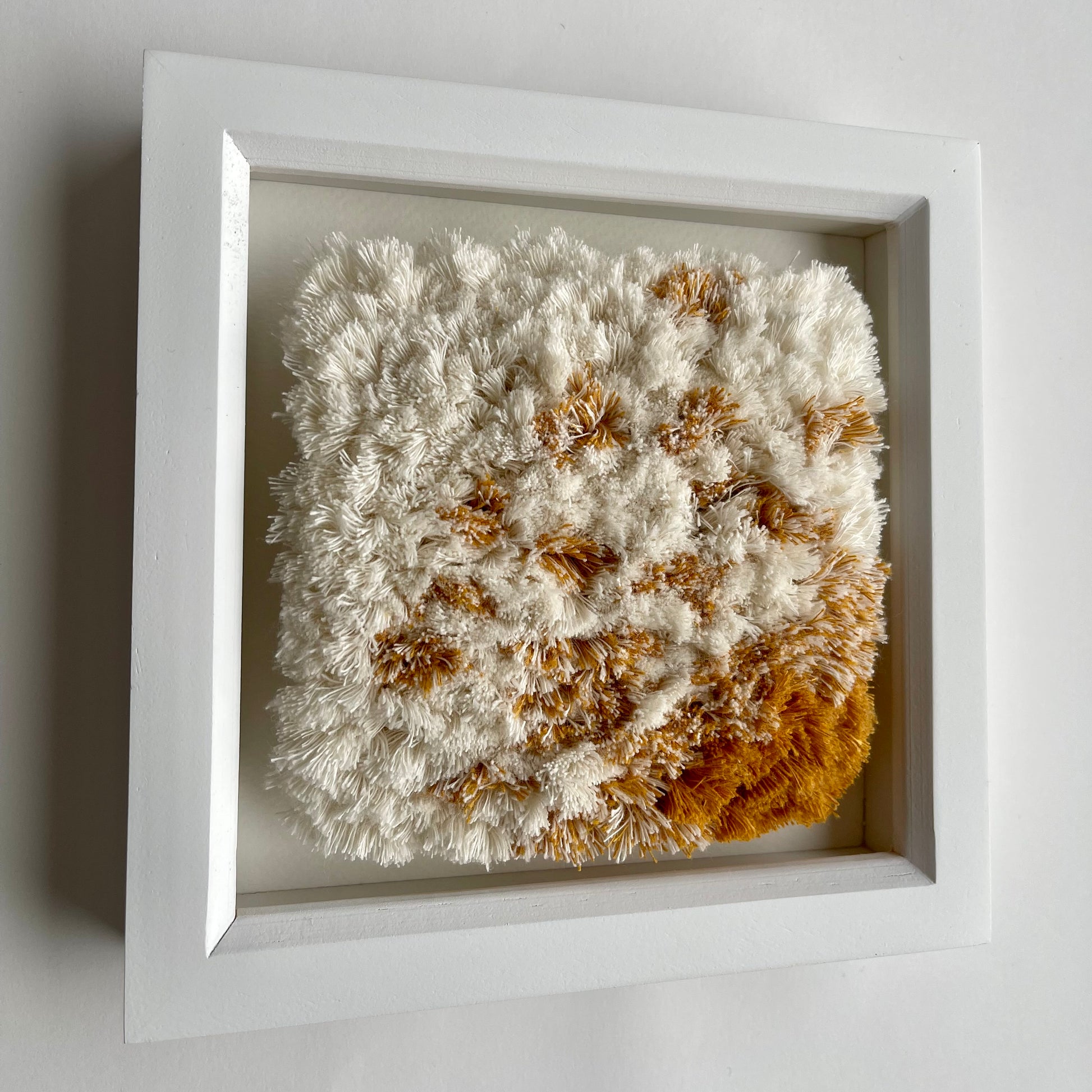 Angled view of cream and yellow ochre fluffy textured woven textile art in square white painted wood  box frame on wall