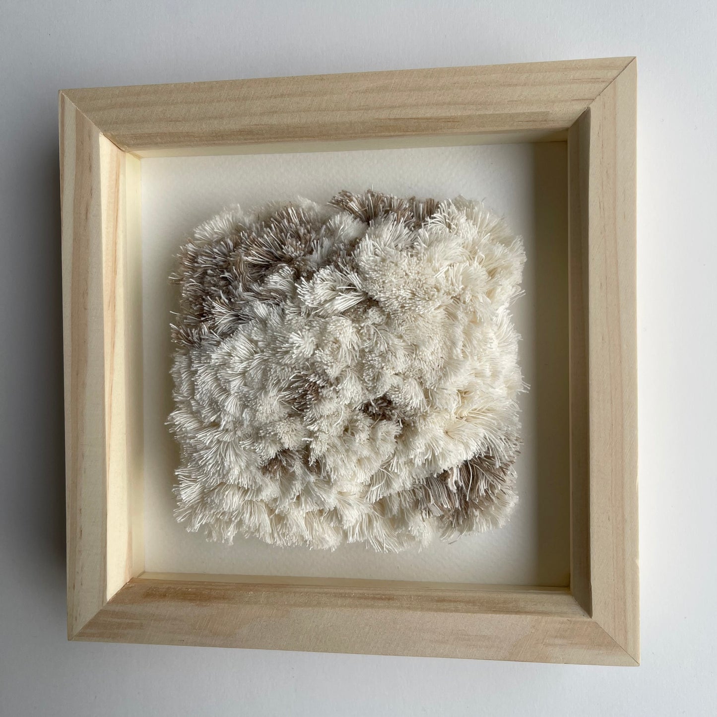 Angled view of cream and beige fluffy textured woven textile art in square natural wood deep box frame on wall