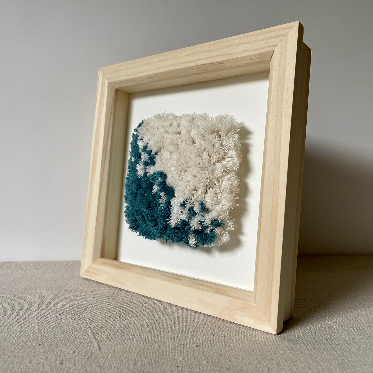 Angled view of teal and cream fluffy textured woven textile art in a square natural wood deep box frame