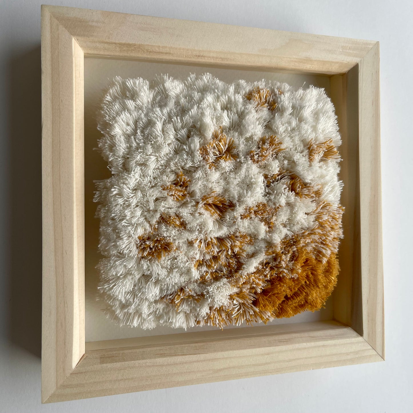 Angled view of cream and yellow ochre fluffy textured woven textile art in natural wood deep box frame on wall