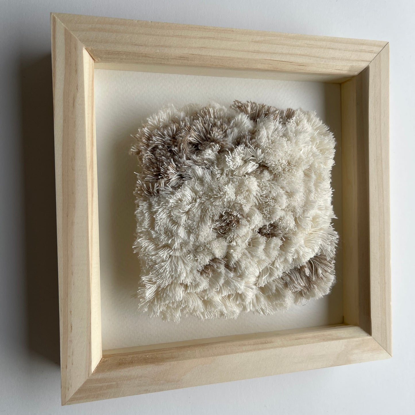 Angled view of cream and beige fluffy textured woven textile art in square natural wood frame on wall