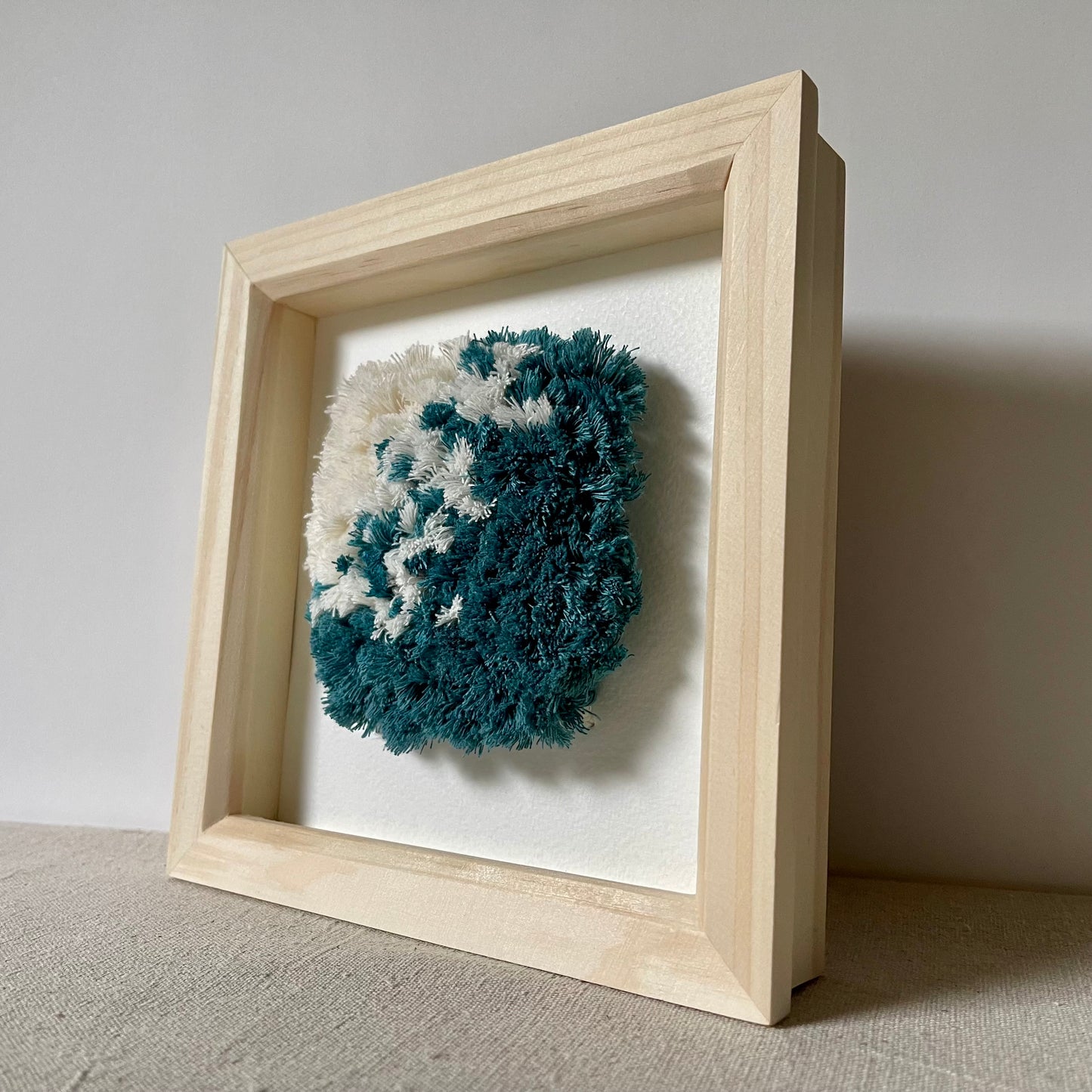 Angled view of ocean teal and cream fluffy textured woven art in a square wood deep box frame