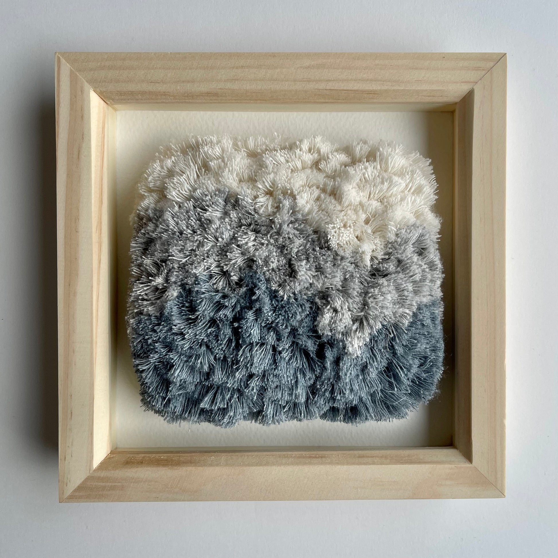 Front view of cream and blue fluffy textured woven textile art in square natural wood deep box frame on wall
