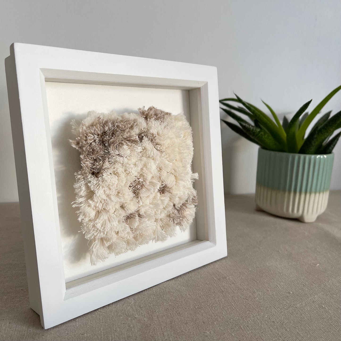 Angled view of cream and beige fluffy textured woven textile art in square white painted wood frame in sunlight