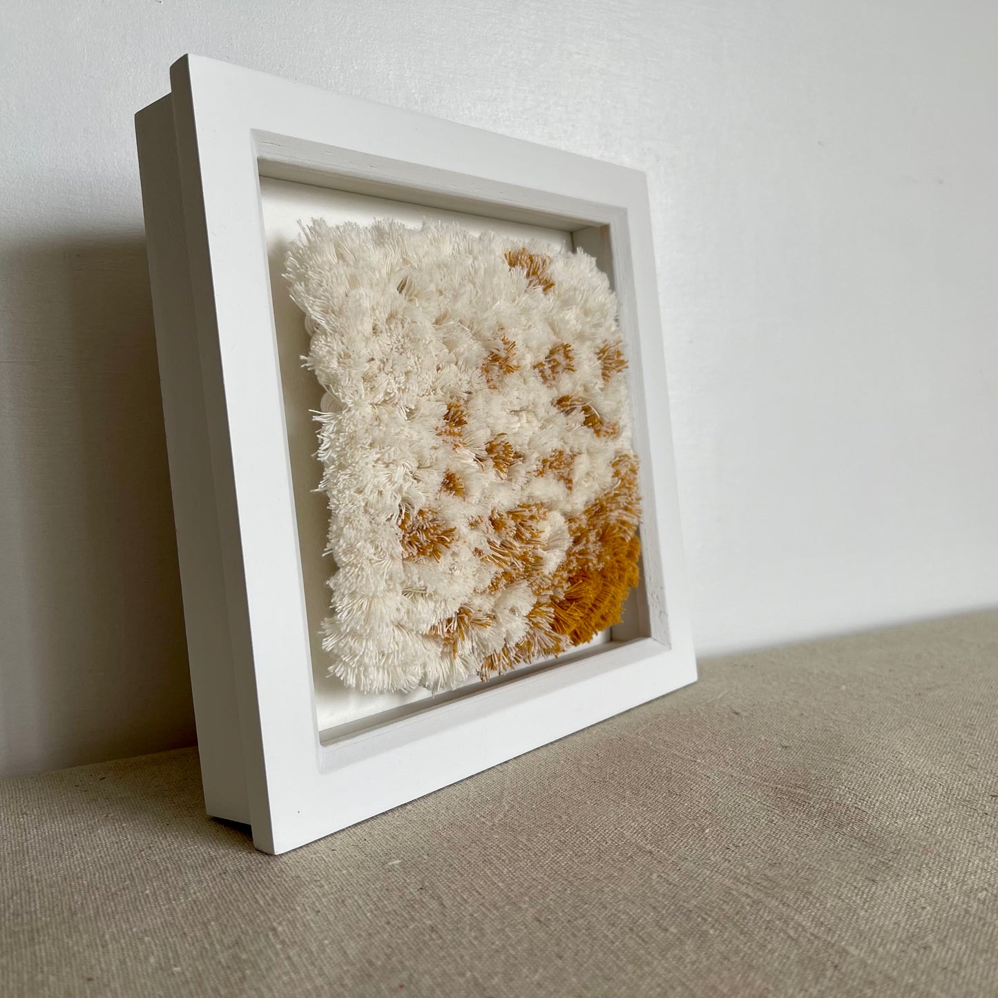 Angled view of cream and yellow ochre fluffy textured woven textile art in square white painted wood deep box frame