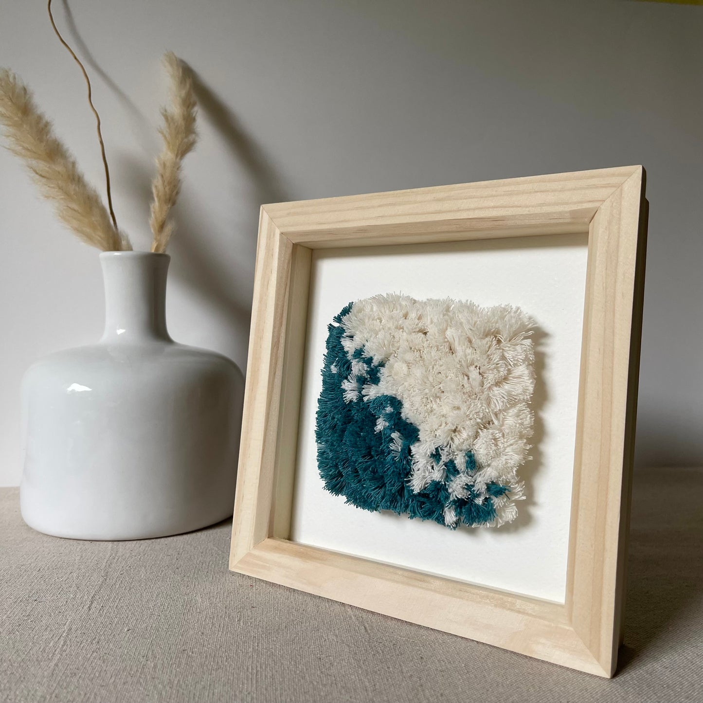 Teal and cream fluffy textured woven textile art in a square natural wood deep box frame next to a scandi style vase