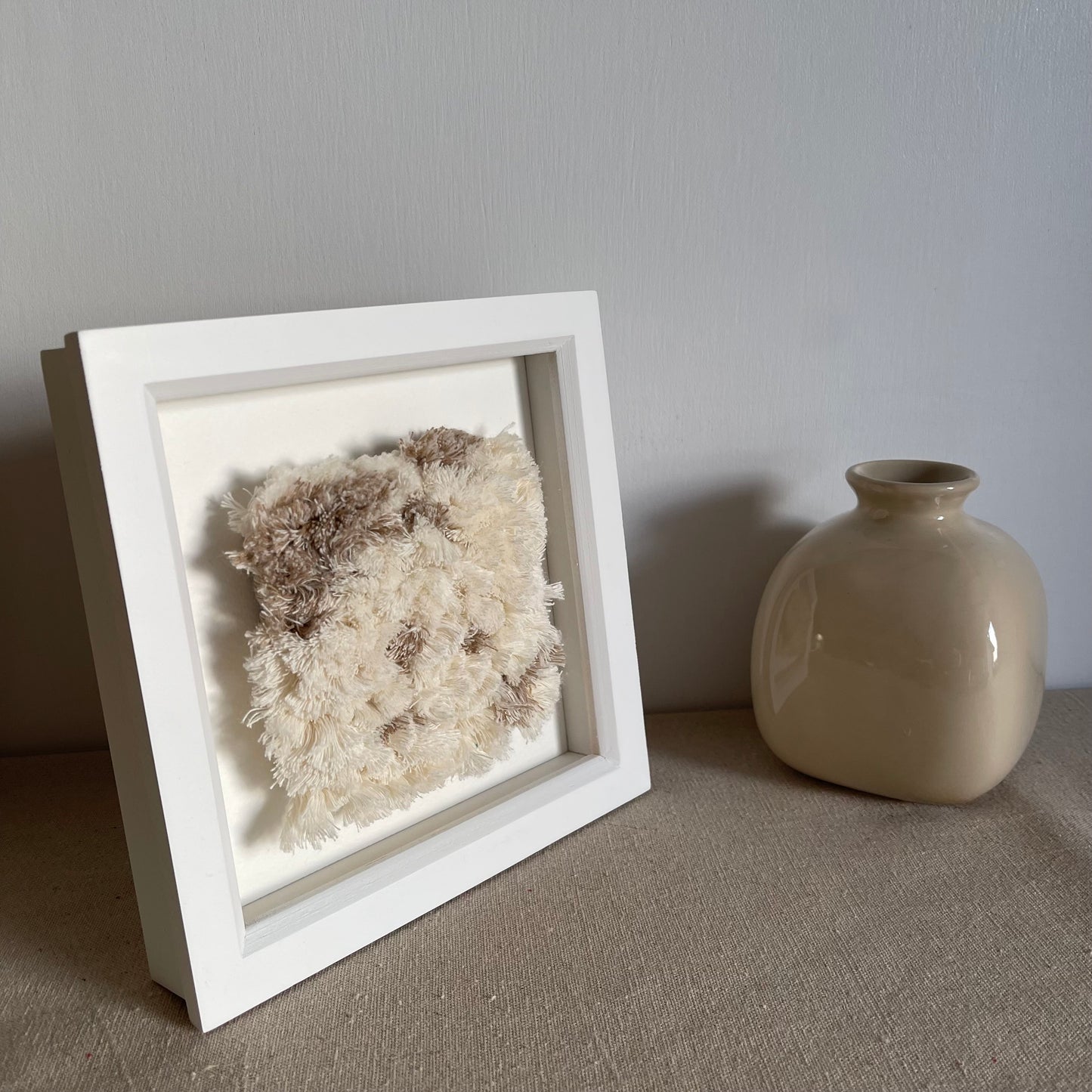 Angled view of cream and beige fluffy textured woven textile art in square white painted wood deep box frame next to scandi style vase