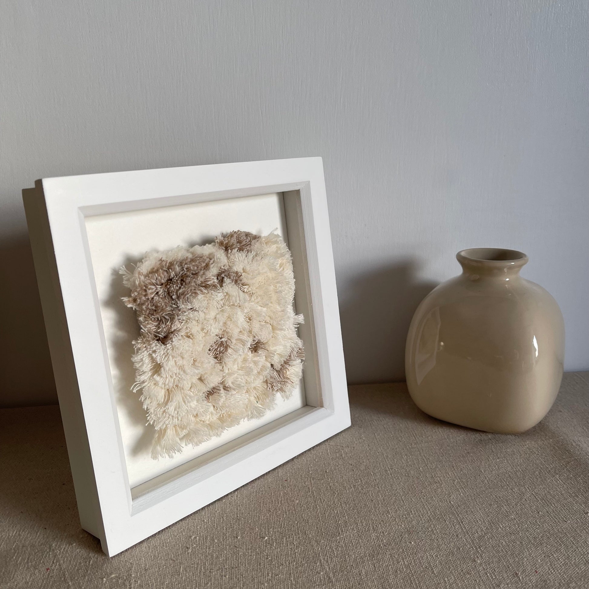 Angled view of cream and beige fluffy textured woven textile art in square white painted wood deep box frame next to scandi style vase
