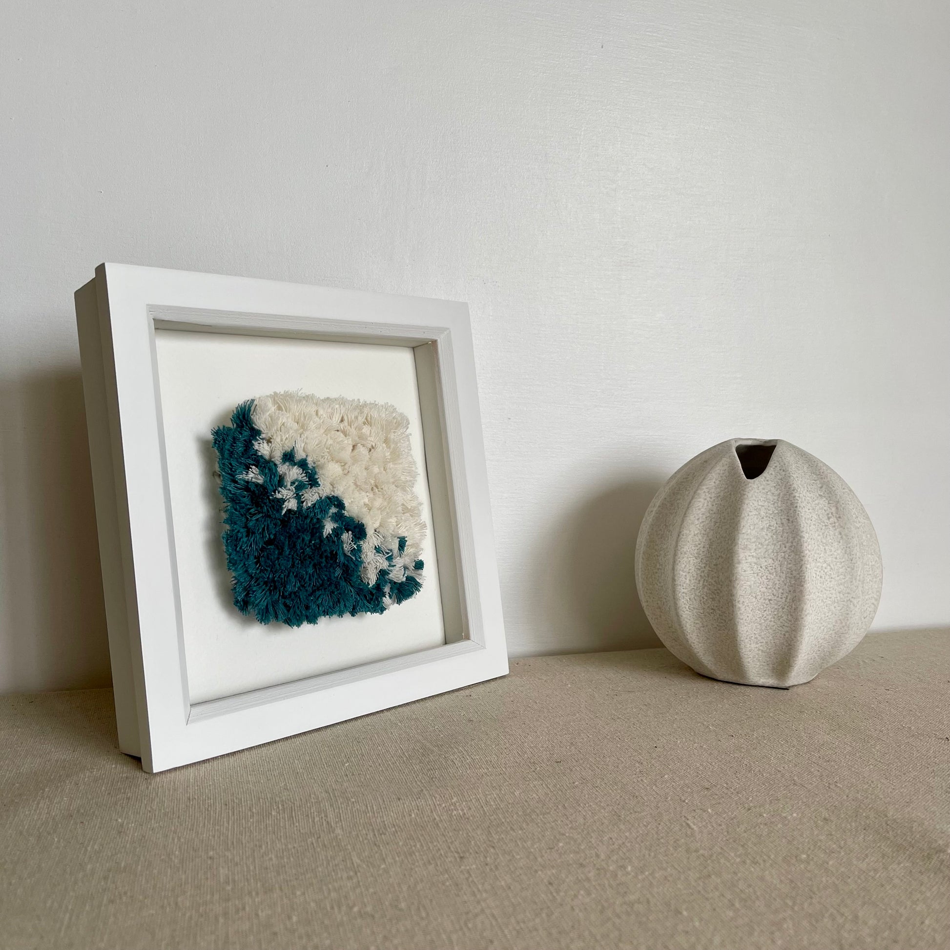 Teal and cream fluffy textured woven textile art in a square white painted wood deep box frame next to a scandi vase