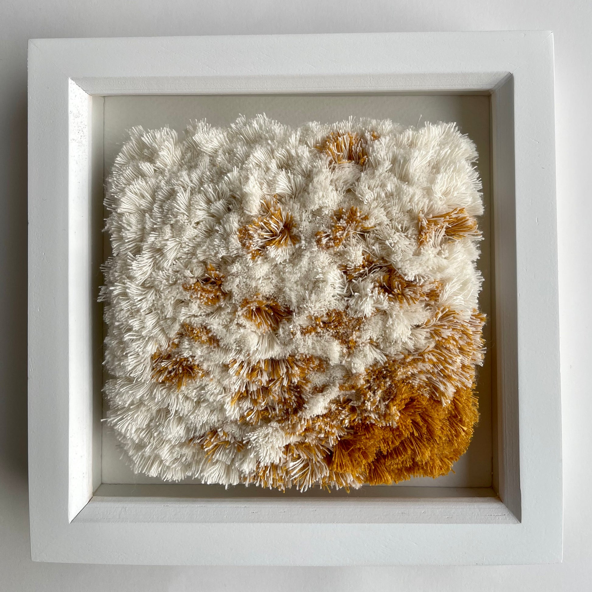 Front view of cream and yellow ochre fluffy textured woven textile art in square white painted wood deep box frame on wall