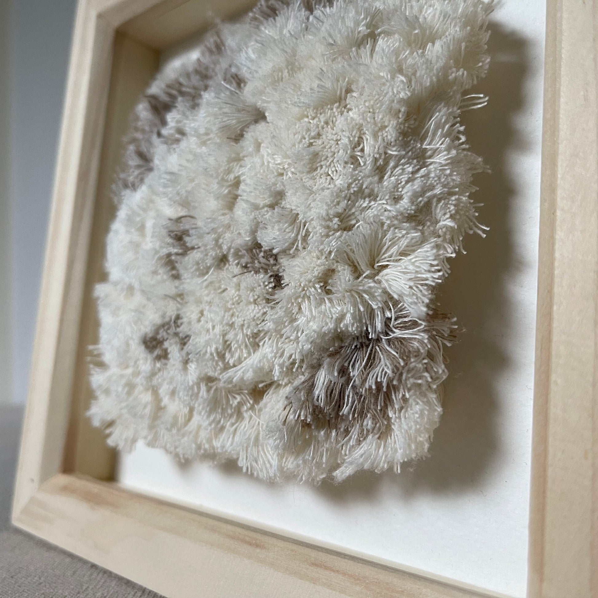 Angled close up detail of cream and beige fluffy textured woven textile art in square natural wood frame