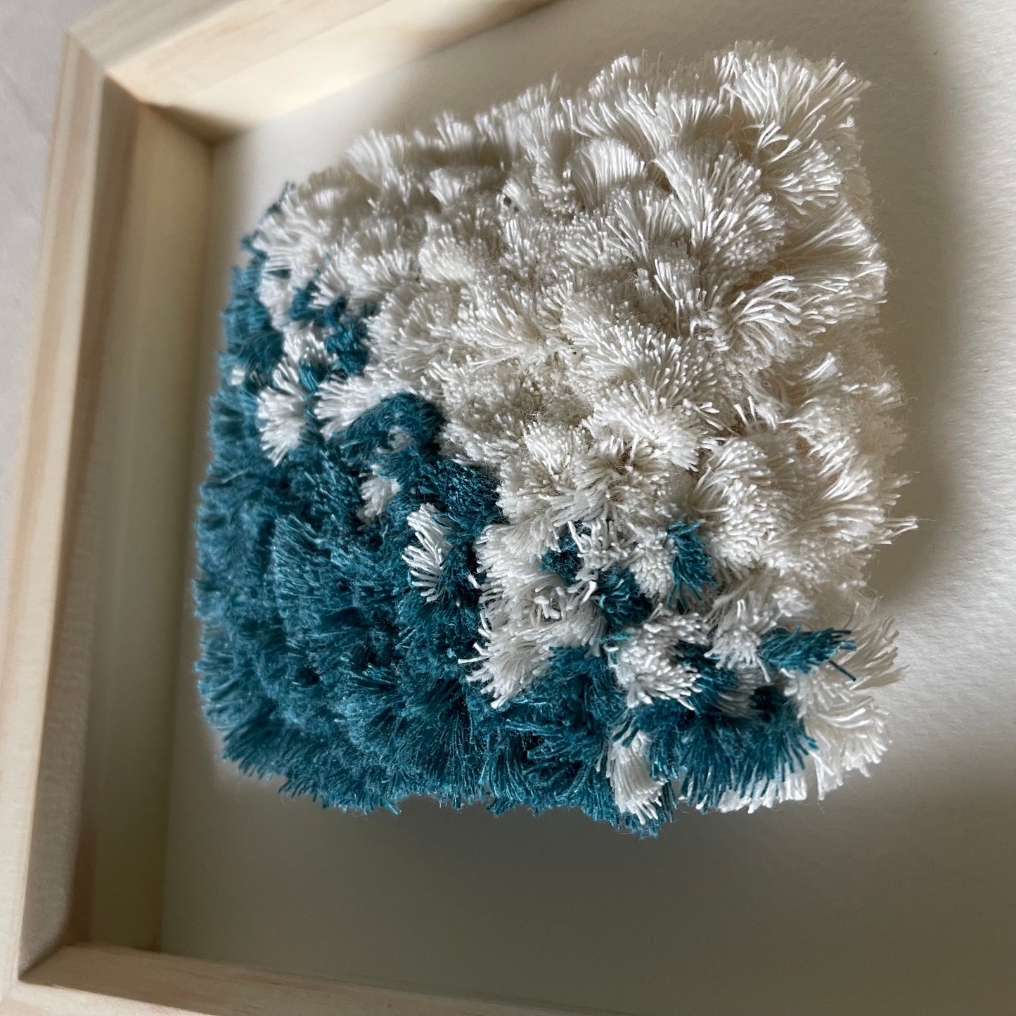 Close up view of teal and cream fluffy woven textile art in a square natural wood deep box frame