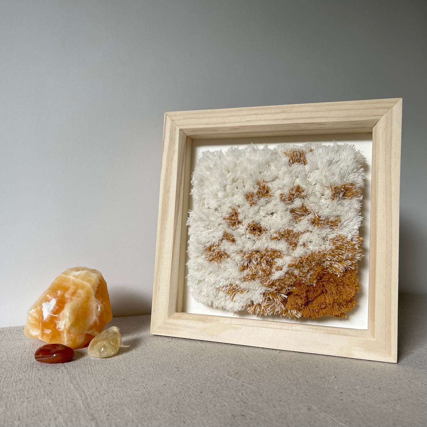 Cream and yellow ochre fluffy textured woven textile art in natural wood deep box frame next to orange calcite, citrine and carnelian crystals