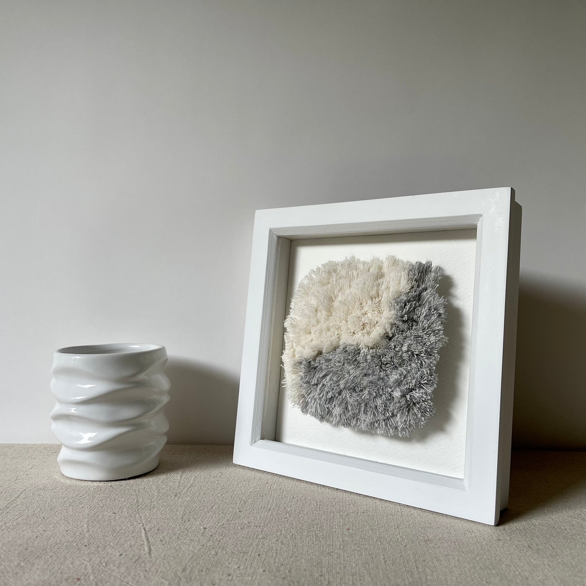 Blue and cream fluffy textured woven textile art in square white painted wood deep box frame next to white ceramic vase