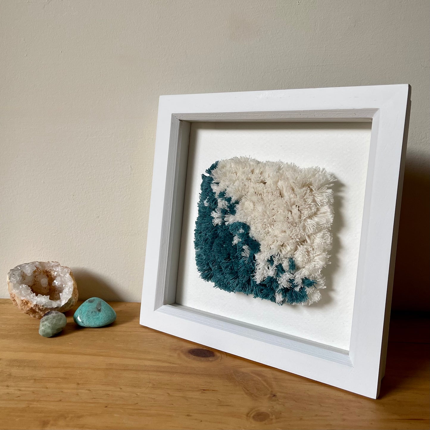 Teal and cream fluffy textured woven textile art in a square white painted wood deep box frame next to aquamarine, fluorite and geode crystals