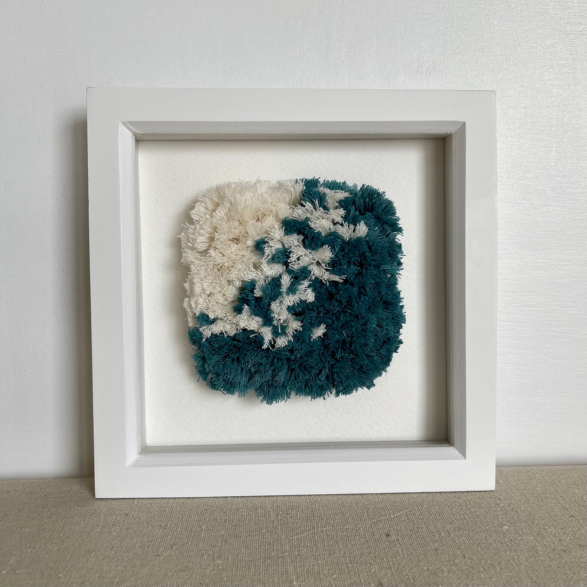 Front view of ocean teal and cream fluffy textured woven art in a square white painted wood deep box frame