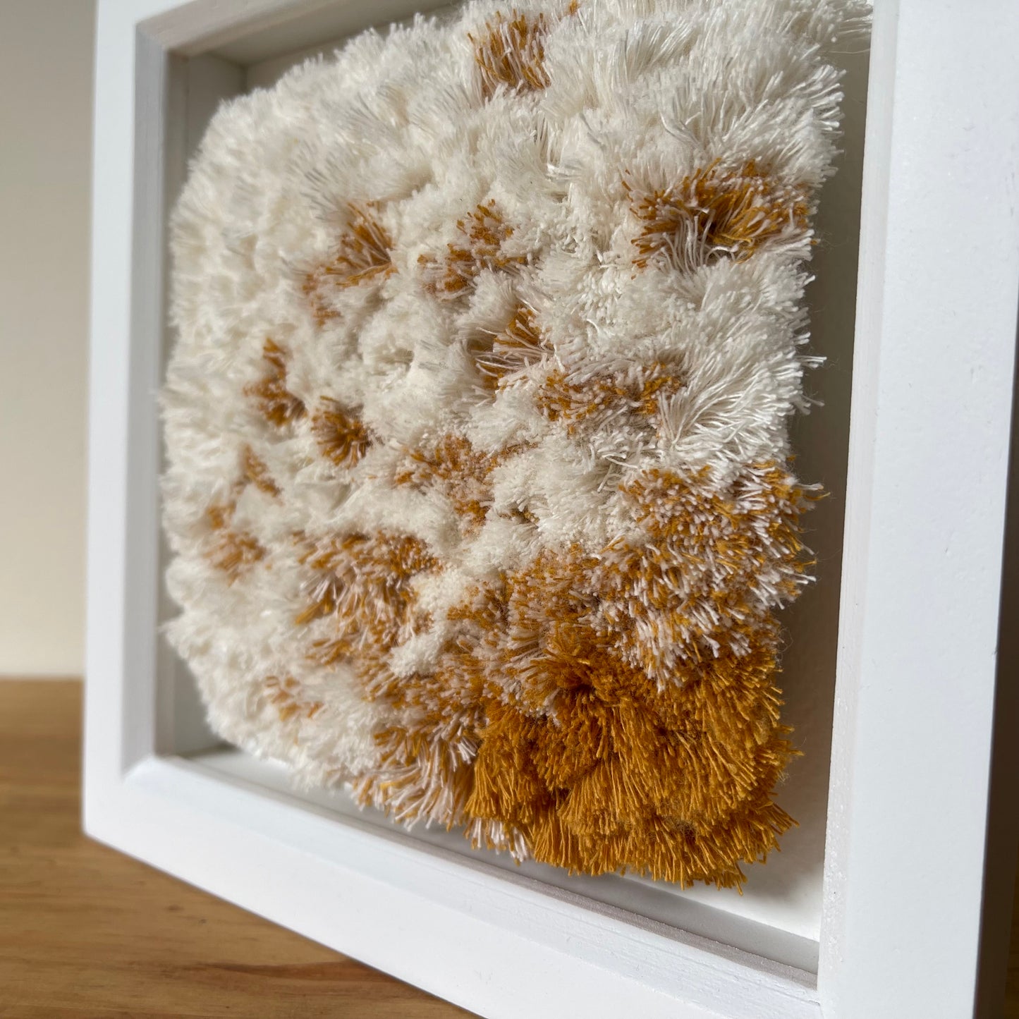 Close up view of cream and yellow ochre fluffy textured woven textile art in square white painted wood deep box frame