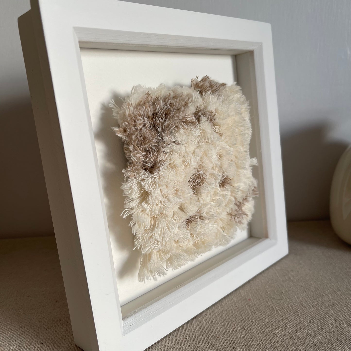 Close up angled view of cream and beige fluffy textured woven textile art in square white painted wood deep box frame