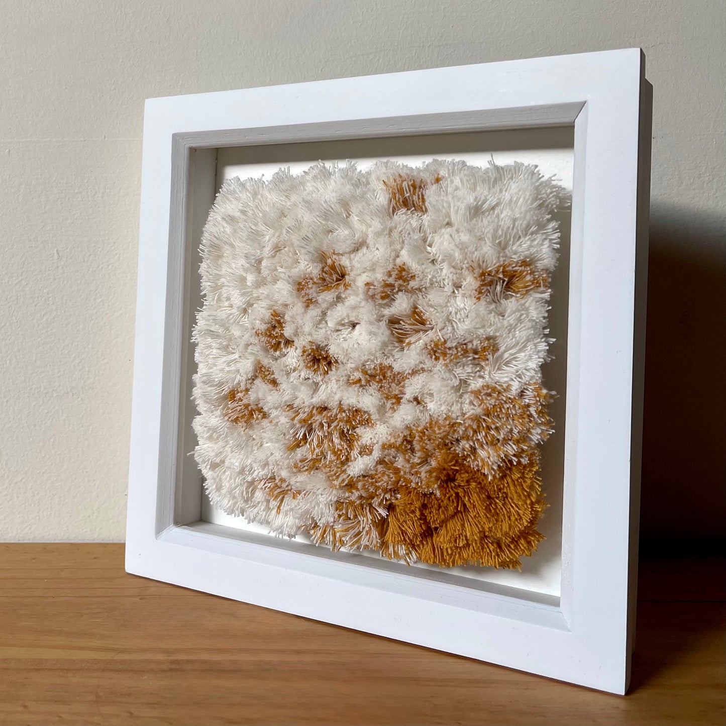 Angled view of cream and yellow ochre fluffy textured woven textile art in square white painted wood  box frame