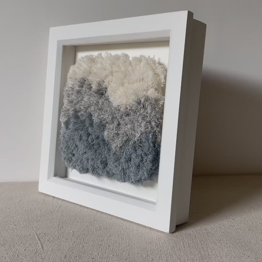 Video of cream and blue fluffy textured woven textile art in square white painted wood deep box frame
