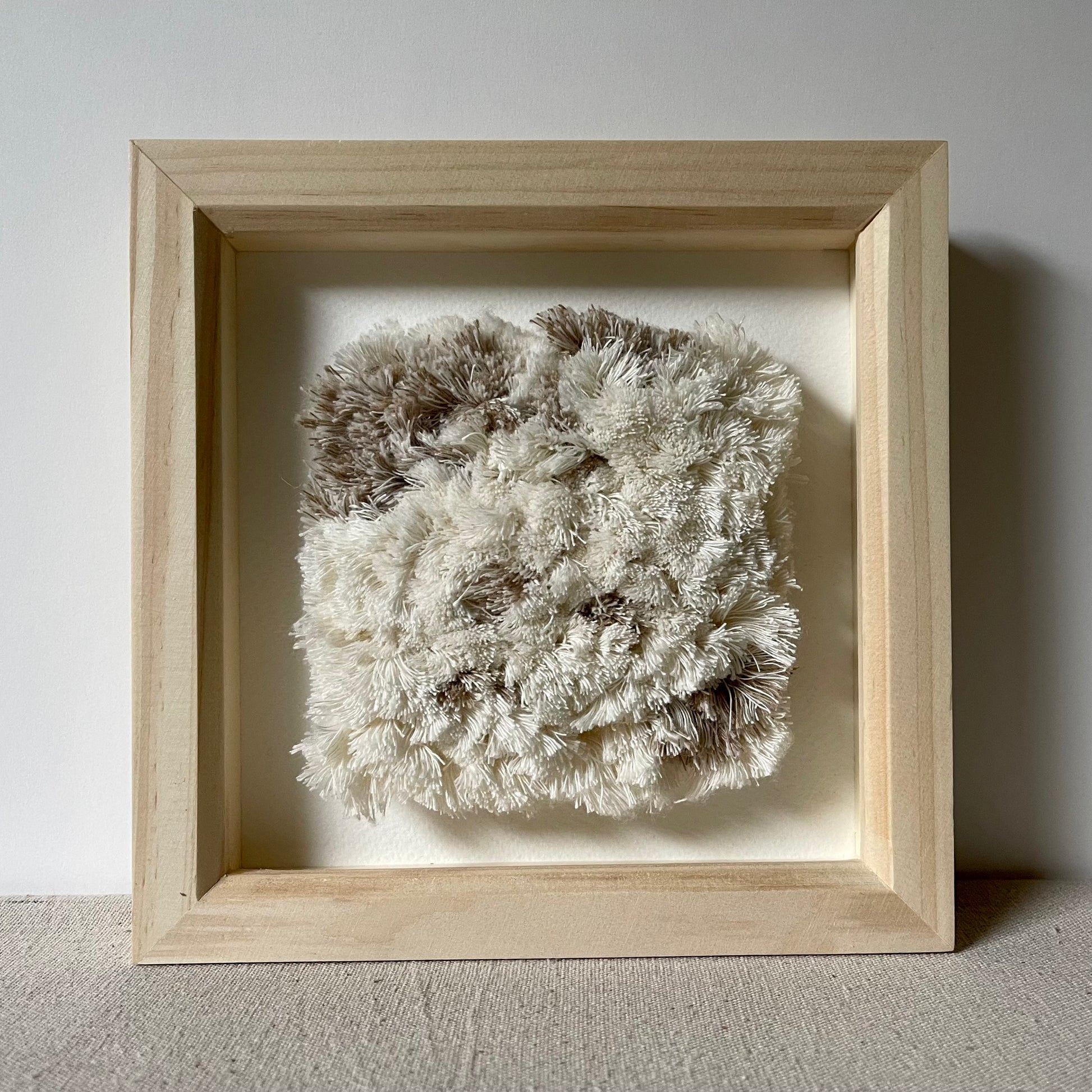 Front view of cream and beige fluffy textured woven textile art in square natural wood deep box frame
