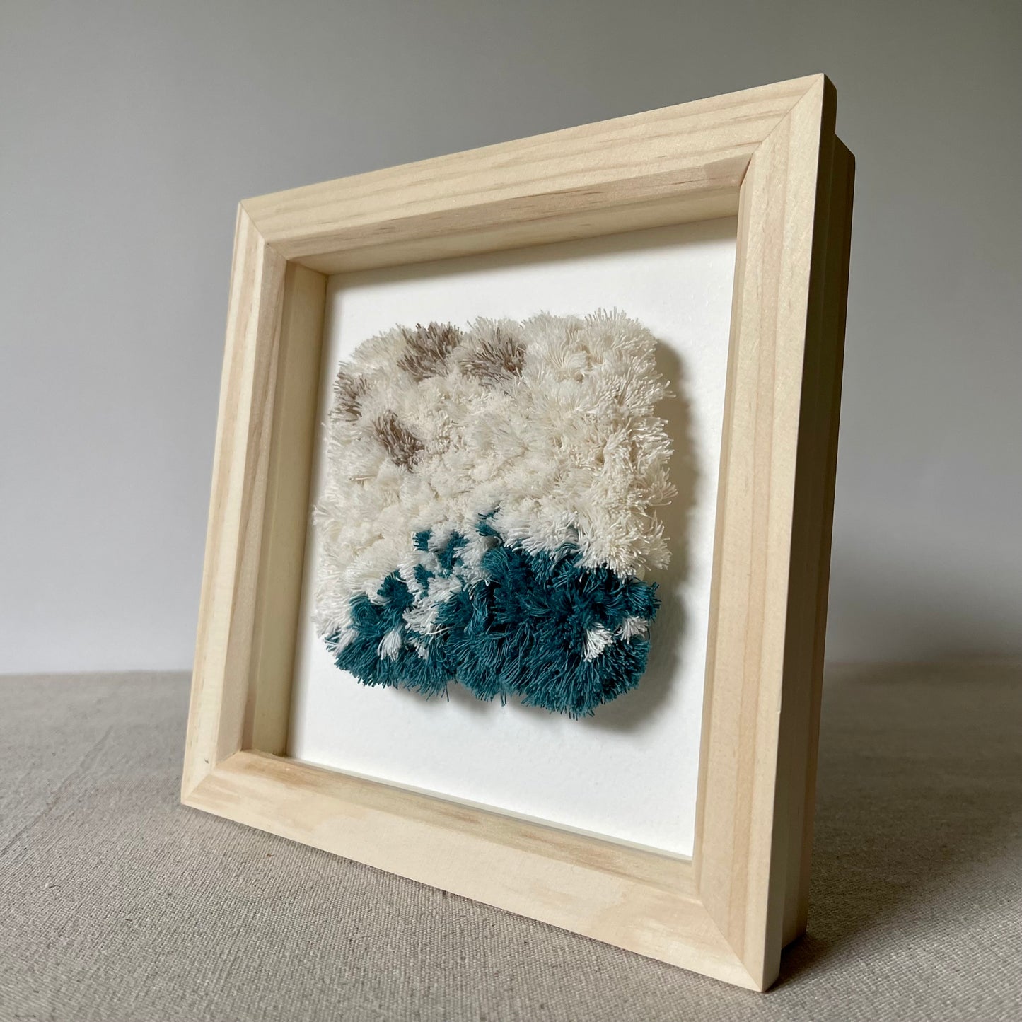 Angled view of cream and ocean teal woven fluffy textured  wall art in a square wood deep box frame