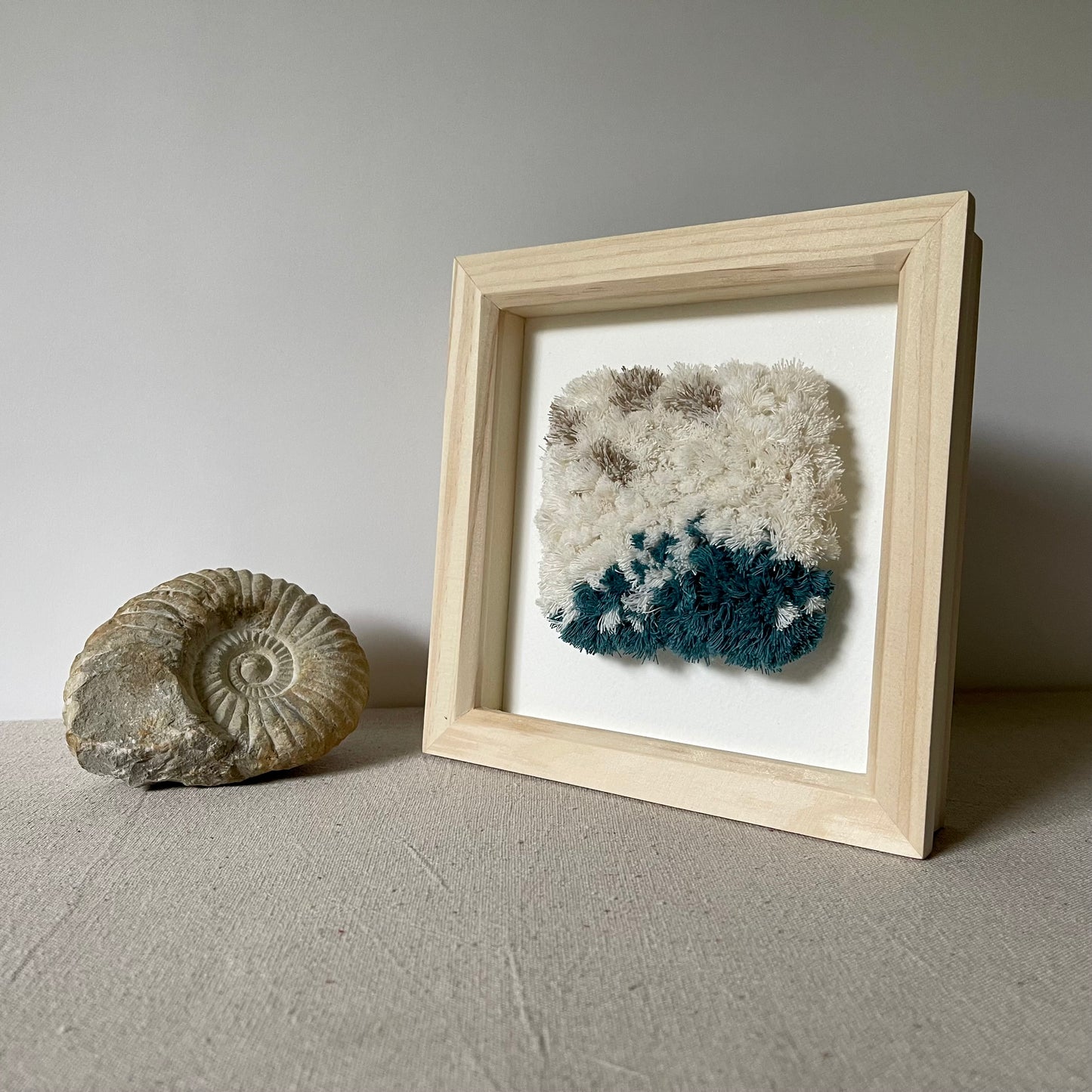Cream and ocean teal woven textured art in a square wood deep box frame next to ammonite