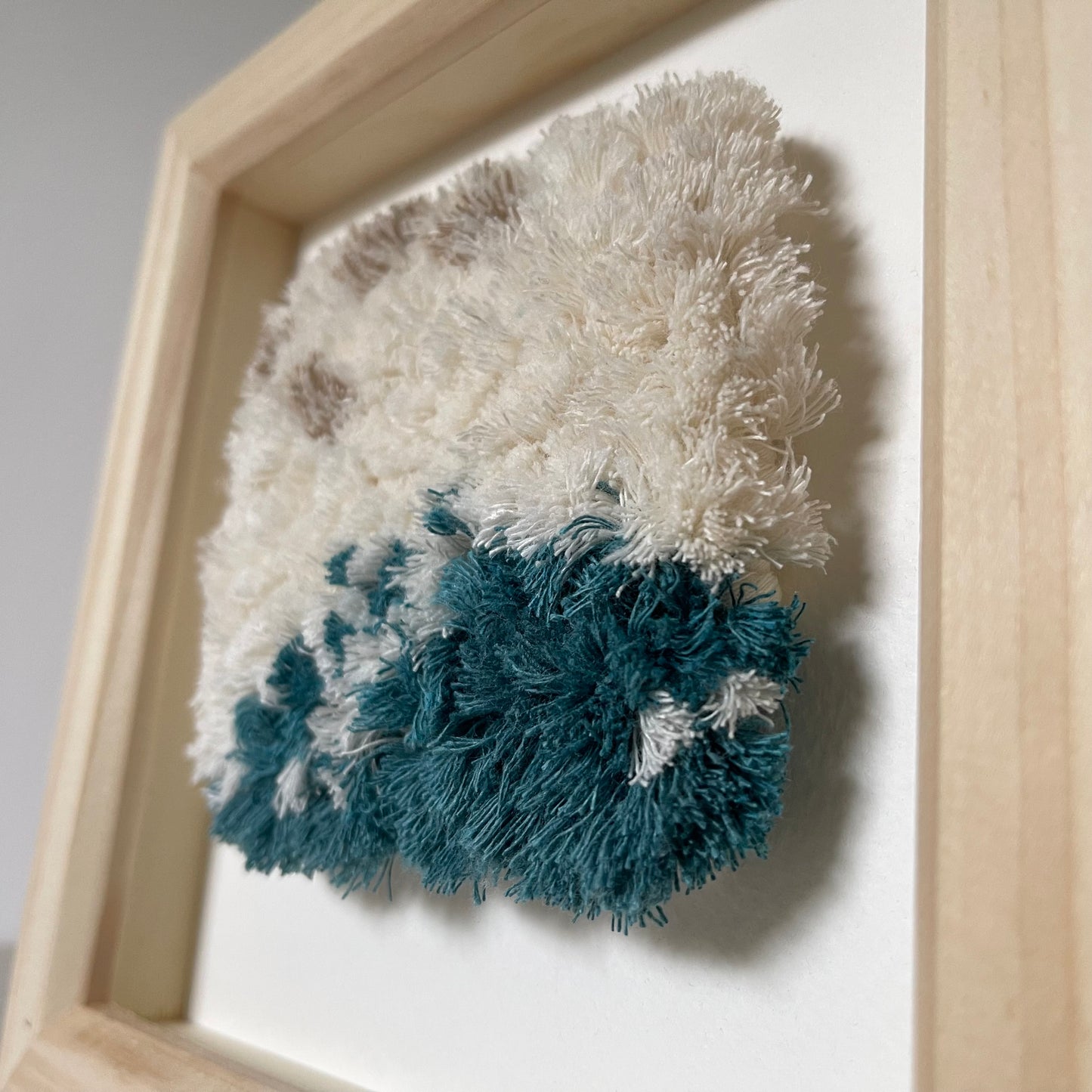 Angled close up of cream and ocean teal woven fluffy textured  art in a square wood frame