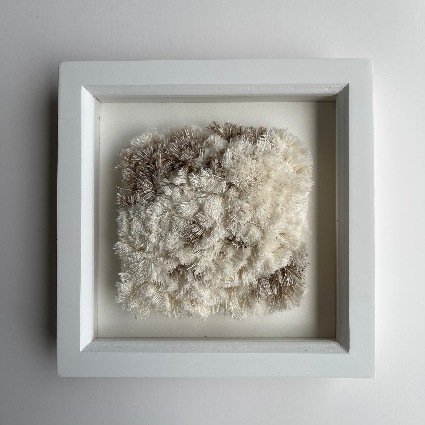Front view of cream and sand textured woven textile art in square white painted wood deep box frame on wal