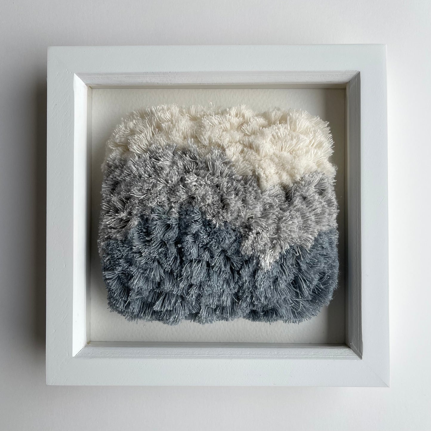 Front view of cream and blue fluffy textured woven textile art in square white painted wood deep box frame on wall