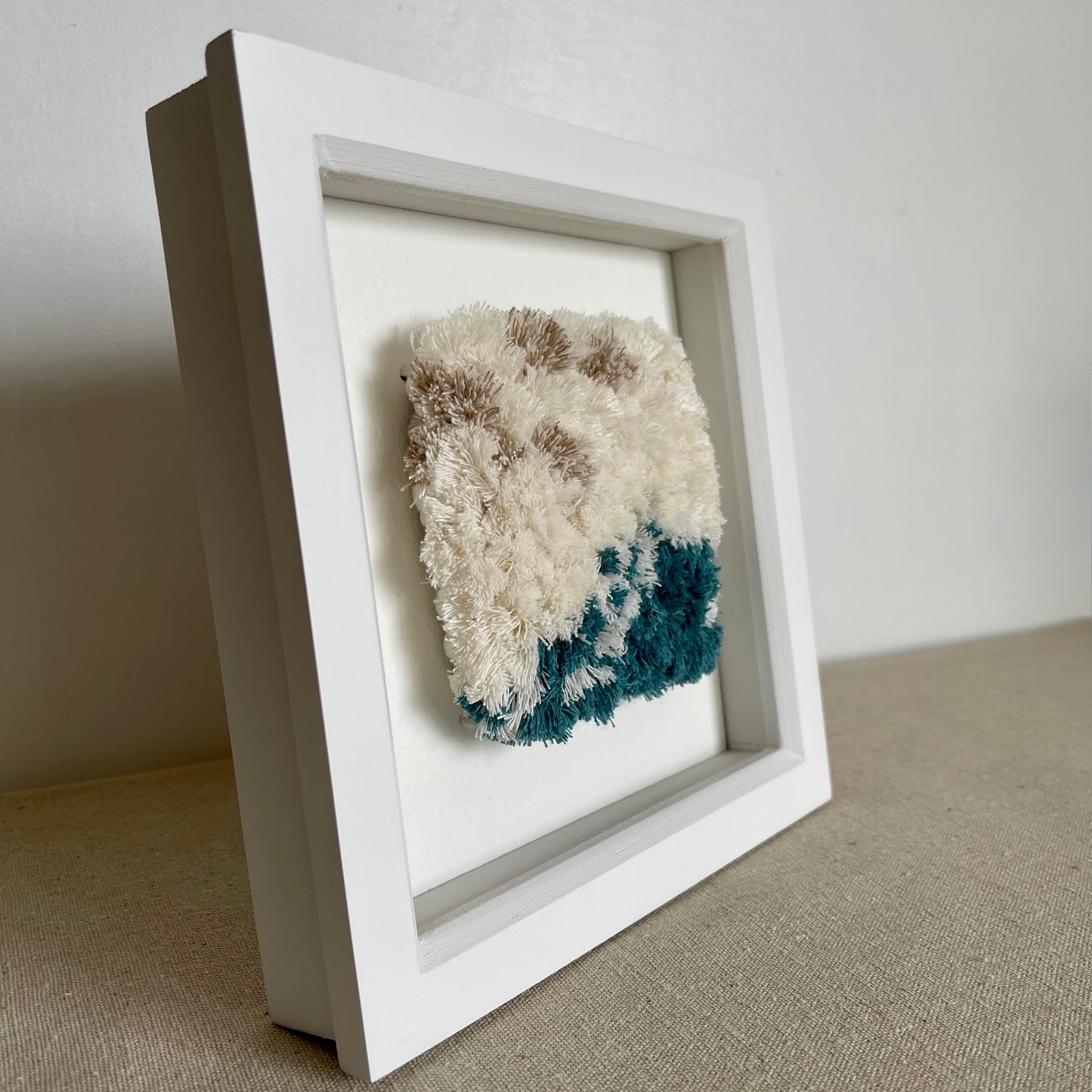 Angled view of cream and ocean teal woven textured art in a square white painted wood frame