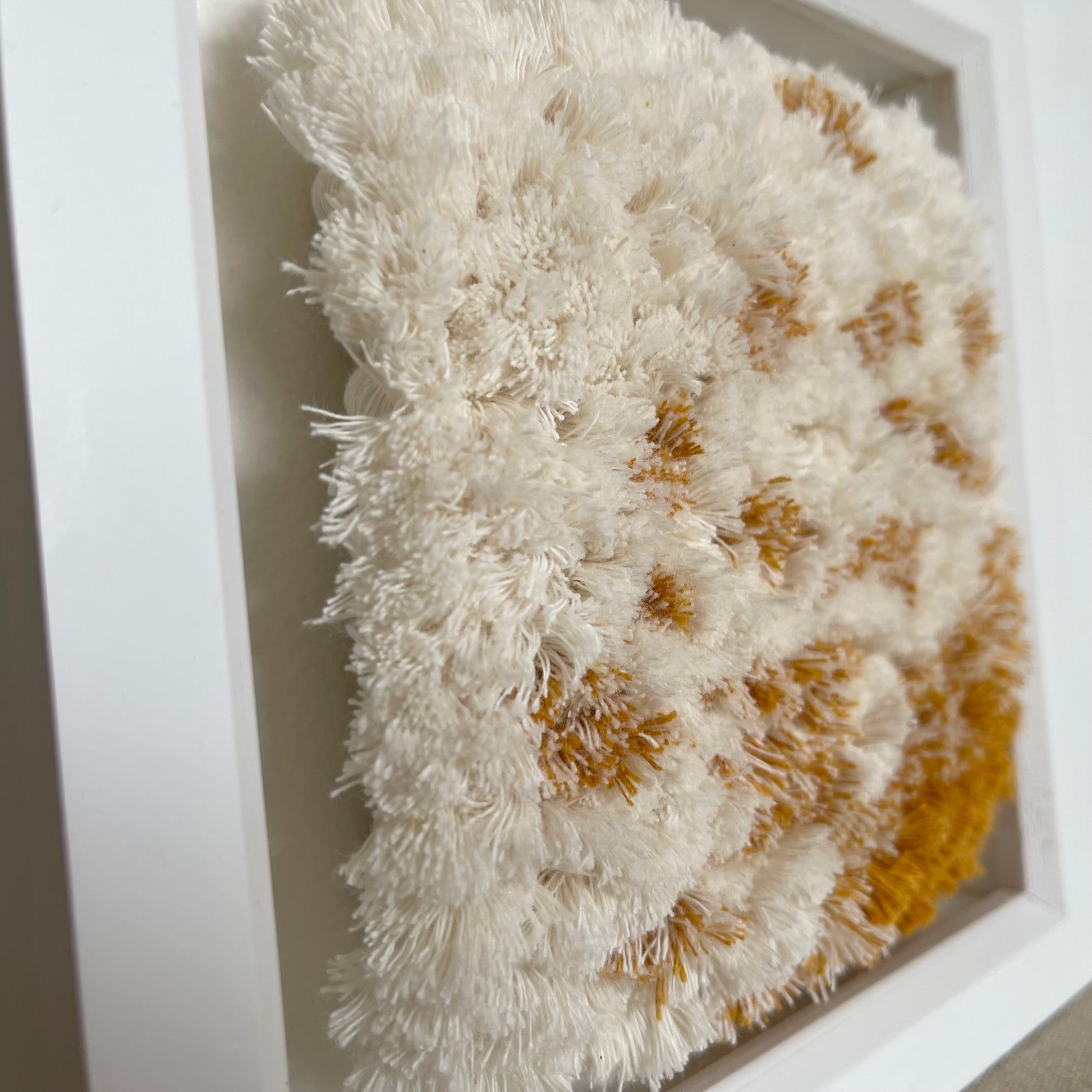 Close up view of cream and yellow ochre fluffy woven textile art in square white painted wood frame